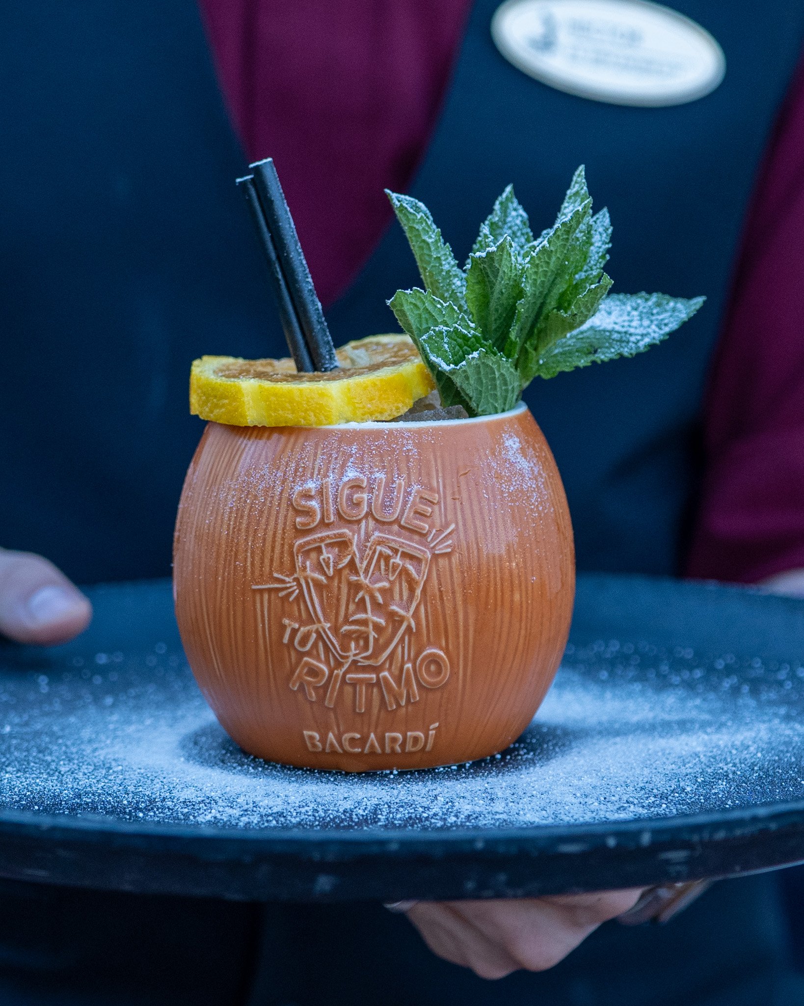 A hand holding a Bacardi glass with a garnish of lemon slice and mint leaves, with a black straw, on a dark surface.