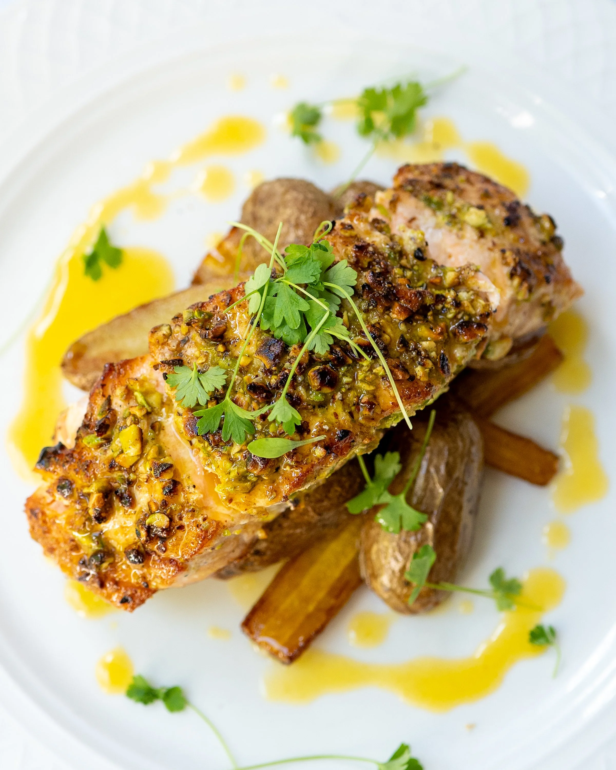Close-up of a piece of grilled fish topped with a pistachio crust, garnished with fresh herbs, served with roasted vegetables and sauce.