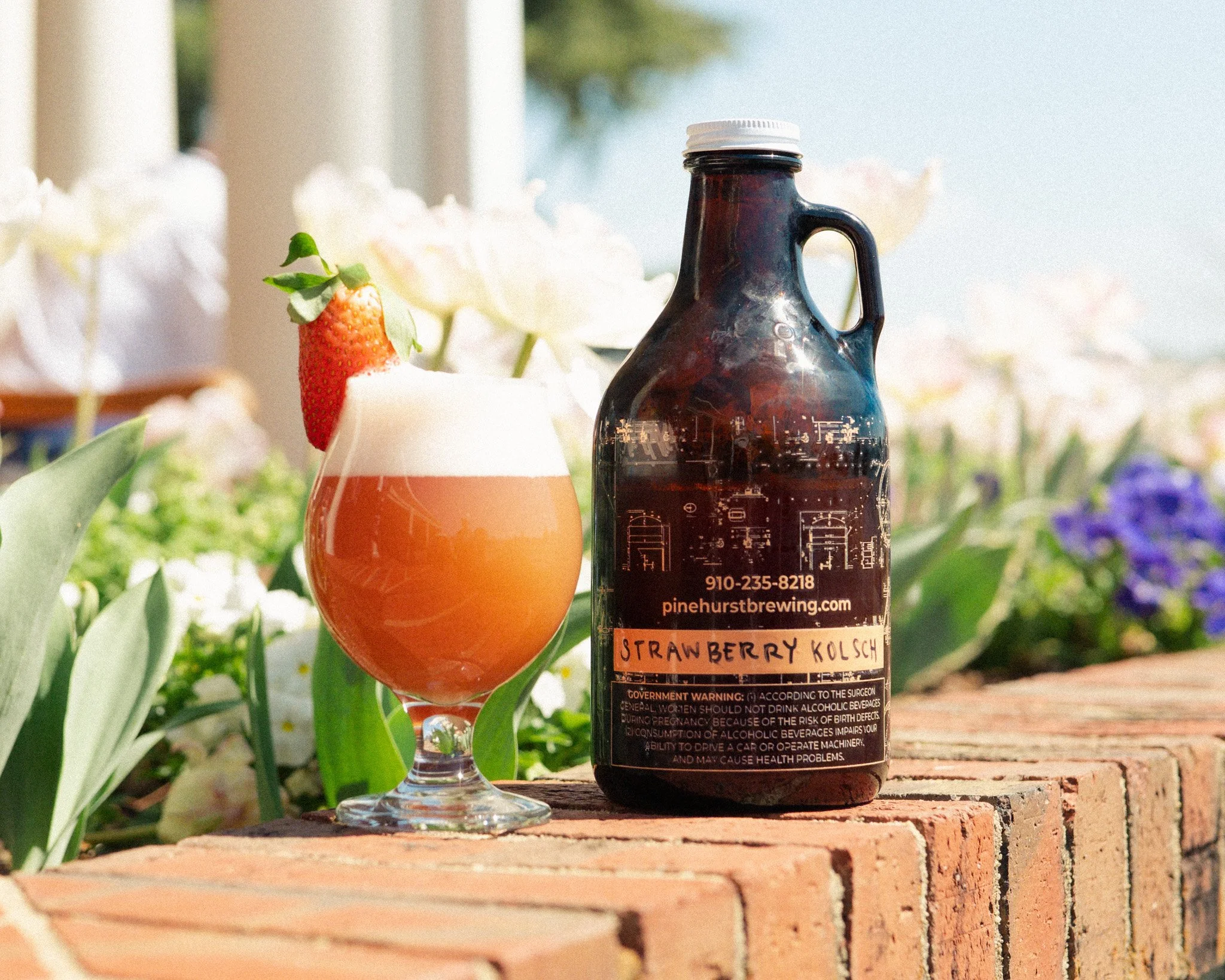 A glass of strawberry kolsch beer with a strawberry garnish next to a bottle of strawberry kolsch beer, both placed on a brick ledge outdoors with green plants and white flowers in the background.
