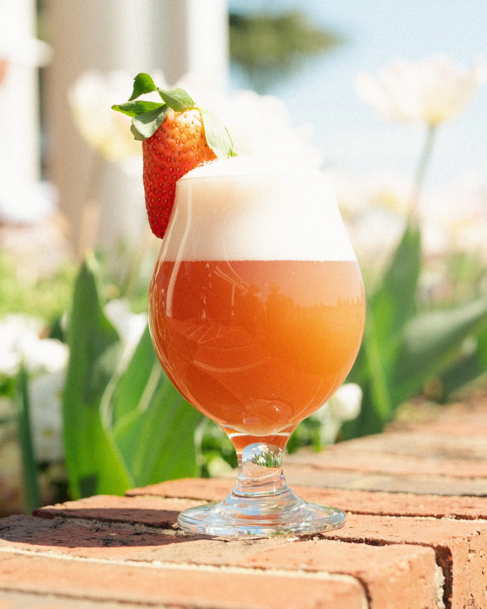 A glass of pink grapefruit juice with a strawberry and mint garnish on top, placed on a brick surface outdoors with greenery in the background.