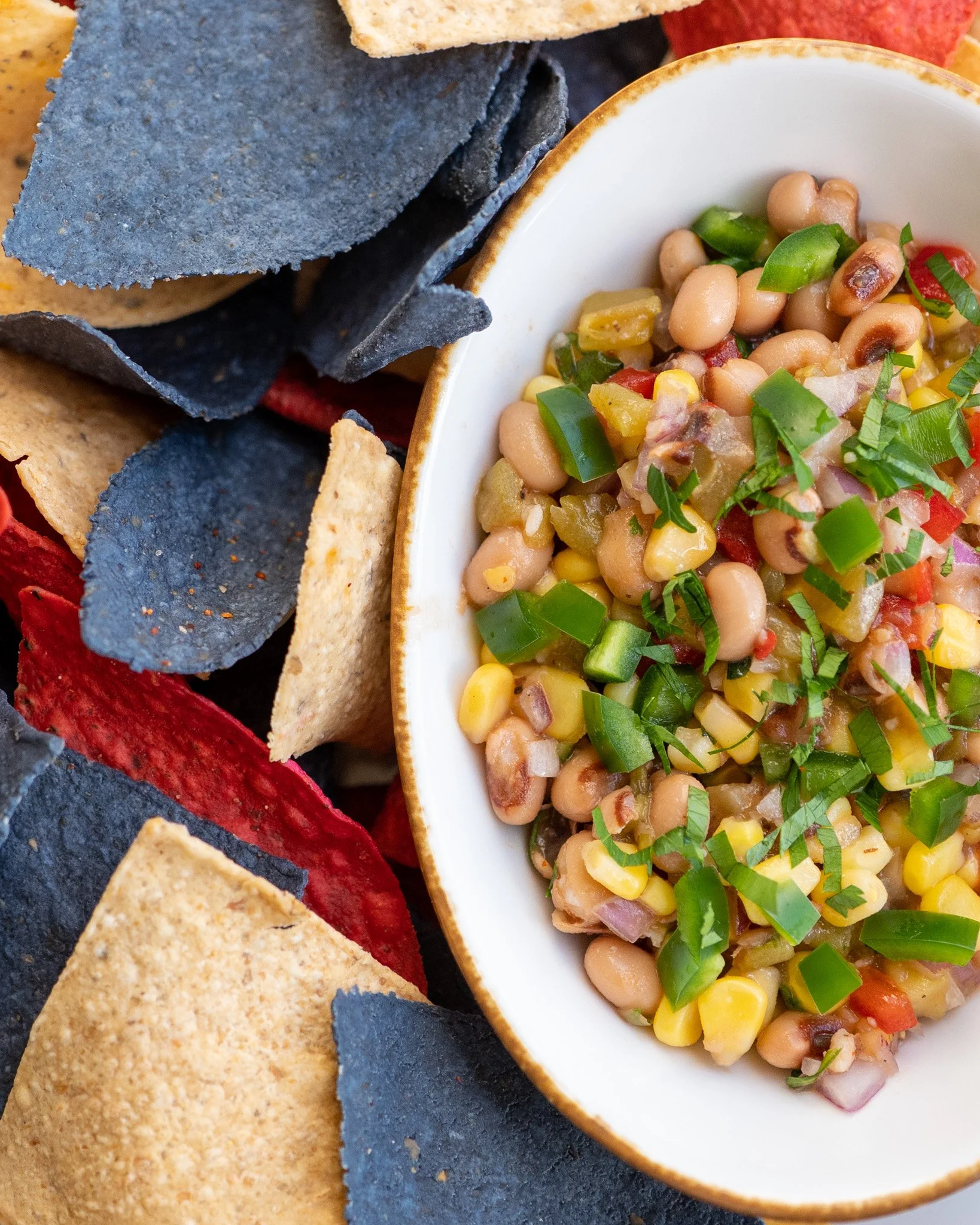 Close-up of a white bowl filled with bean salad garnished with chopped green herbs, surrounded by multicolored tortilla chips.