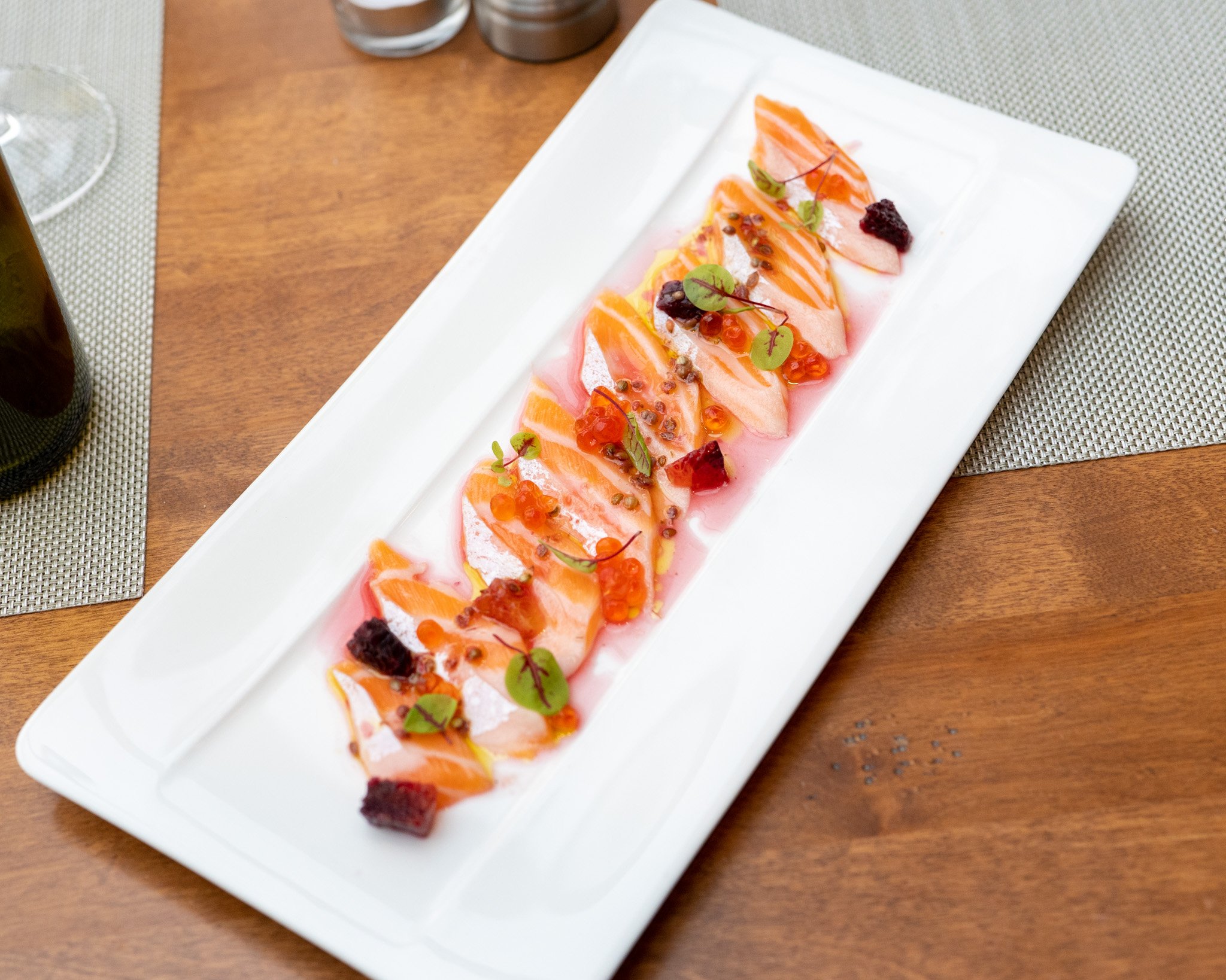 A long rectangular white plate with thin slices of salmon garnished with microgreens, orange sauce, small red spheres, and black crumbs. The plate is on a wooden table with a beige placemat, with glasses and salt and pepper shakers above.