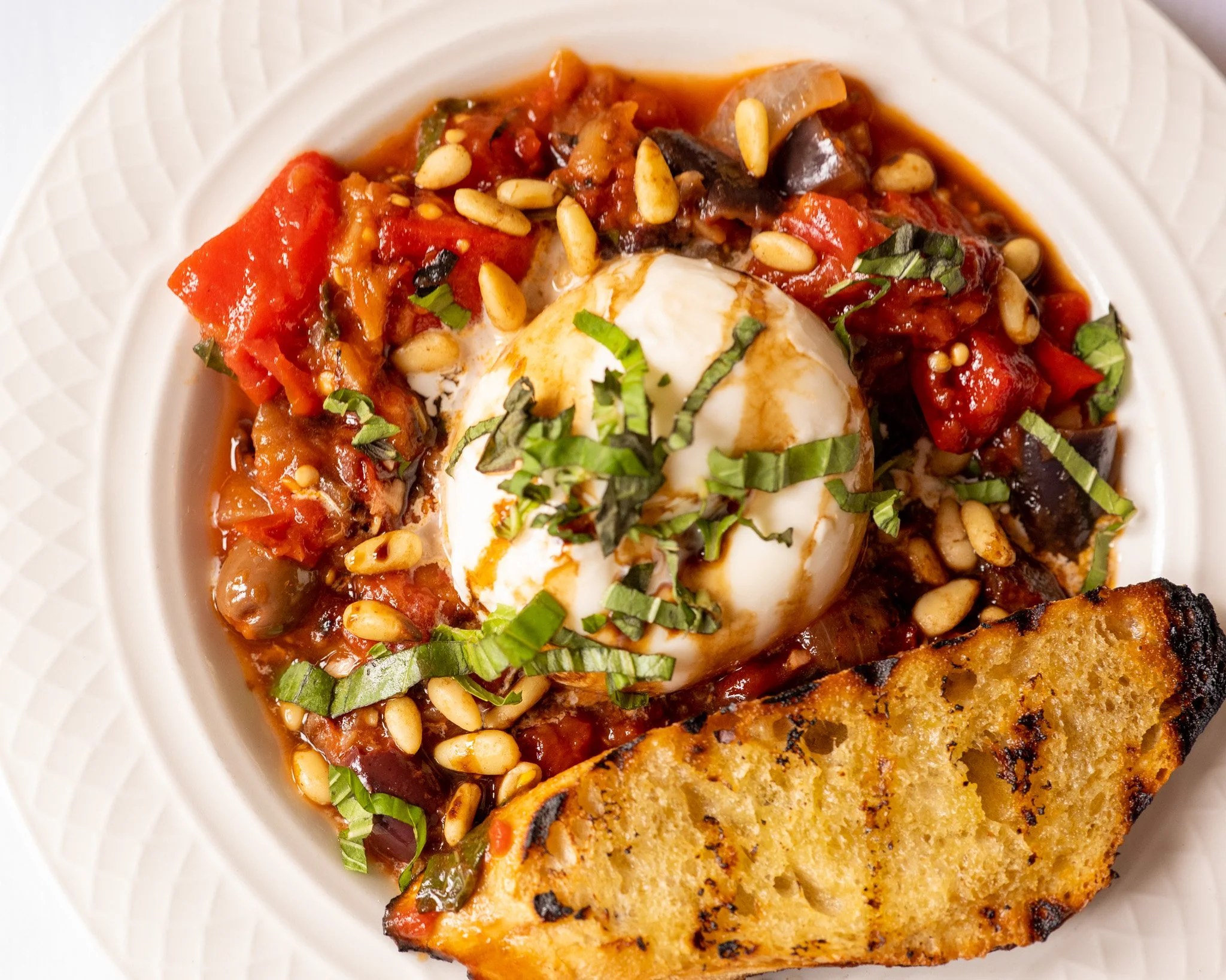 Plate of Italian baked salmon served on a white textured plate, topped with tomato sauce, pine nuts, chopped basil, and a poached egg, with a piece of grilled salmon fillet on the side.