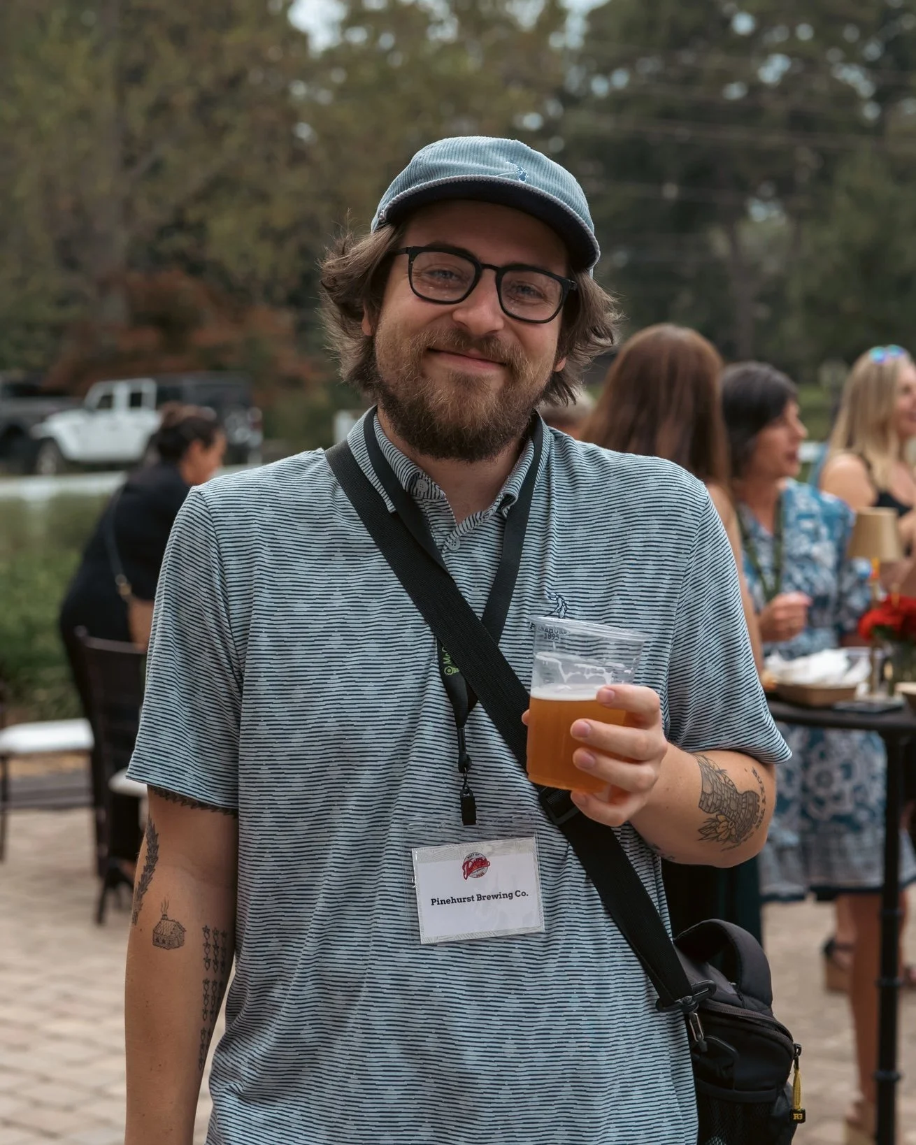 A man with glasses and a beard smiling while holding a plastic cup of beer at an outdoor event. He has tattoos on his arm and is wearing a striped shirt, a light blue cap, and a lanyard with a name tag that reads "Pinehurst Brewing Co." In the background, other people are mingling and a line of parked cars is visible.