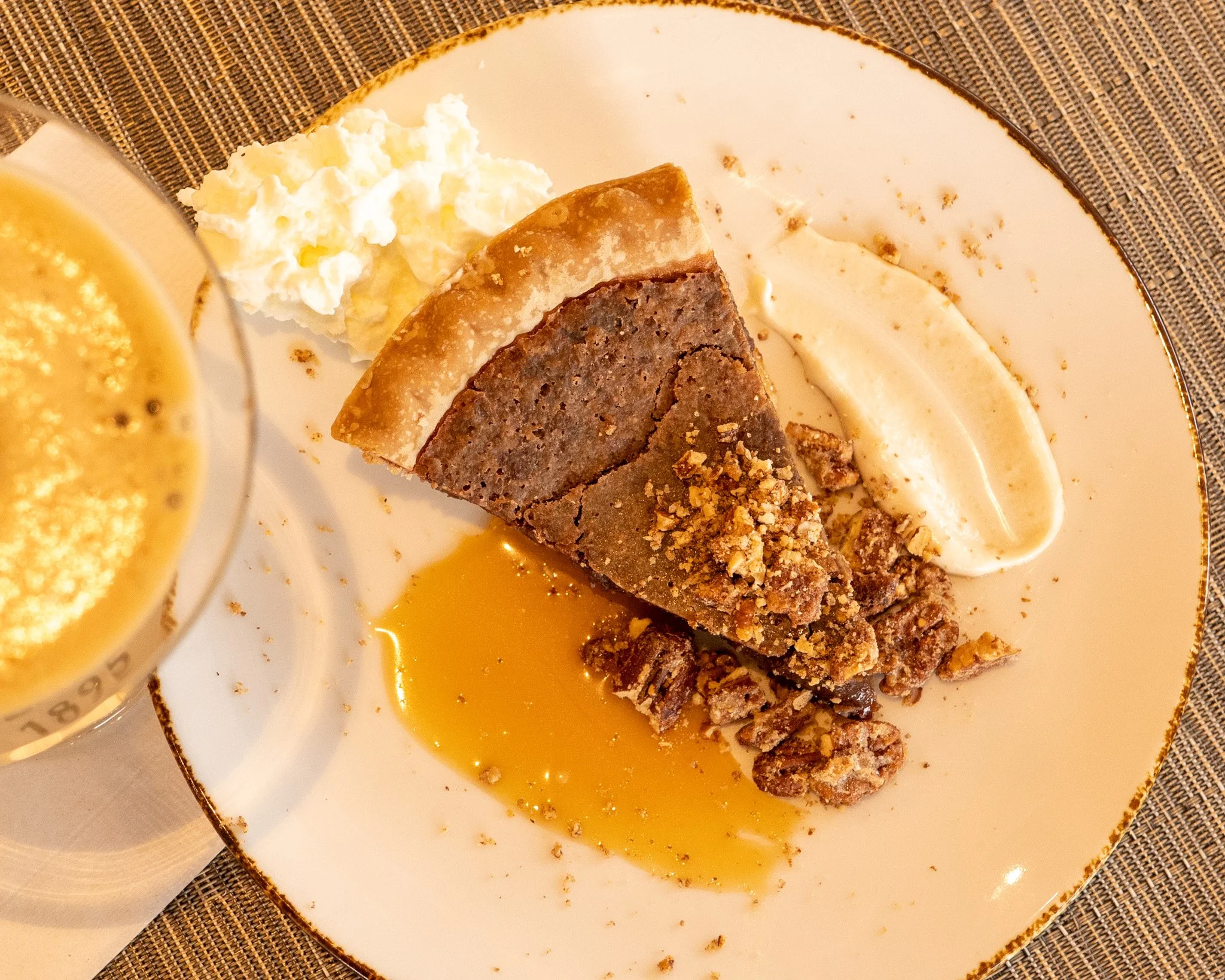 A slice of chocolate cake with crumbs on top, a dollop of whipped cream, and caramel sauce on a white plate with a gold rim, placed on a woven placemat. Part of a glass with a frothy beverage is visible in the foreground.