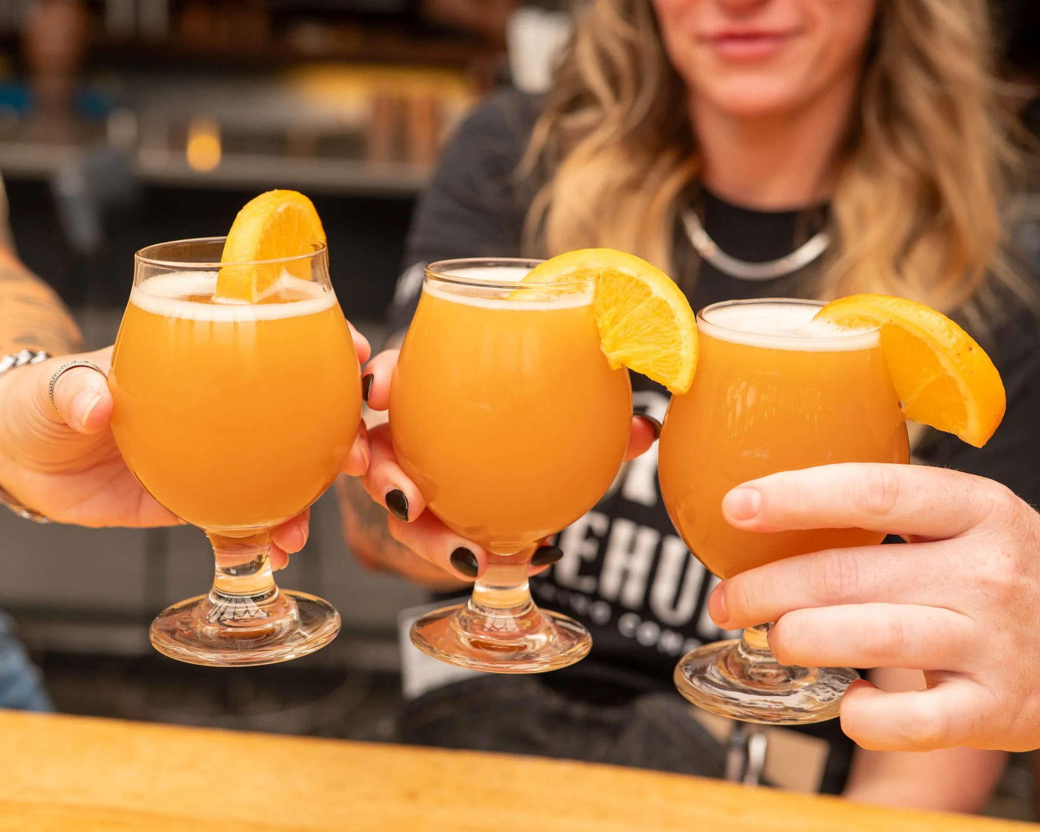 Four glasses of orange-colored beverage with orange slices on top, held by different women in a social setting.