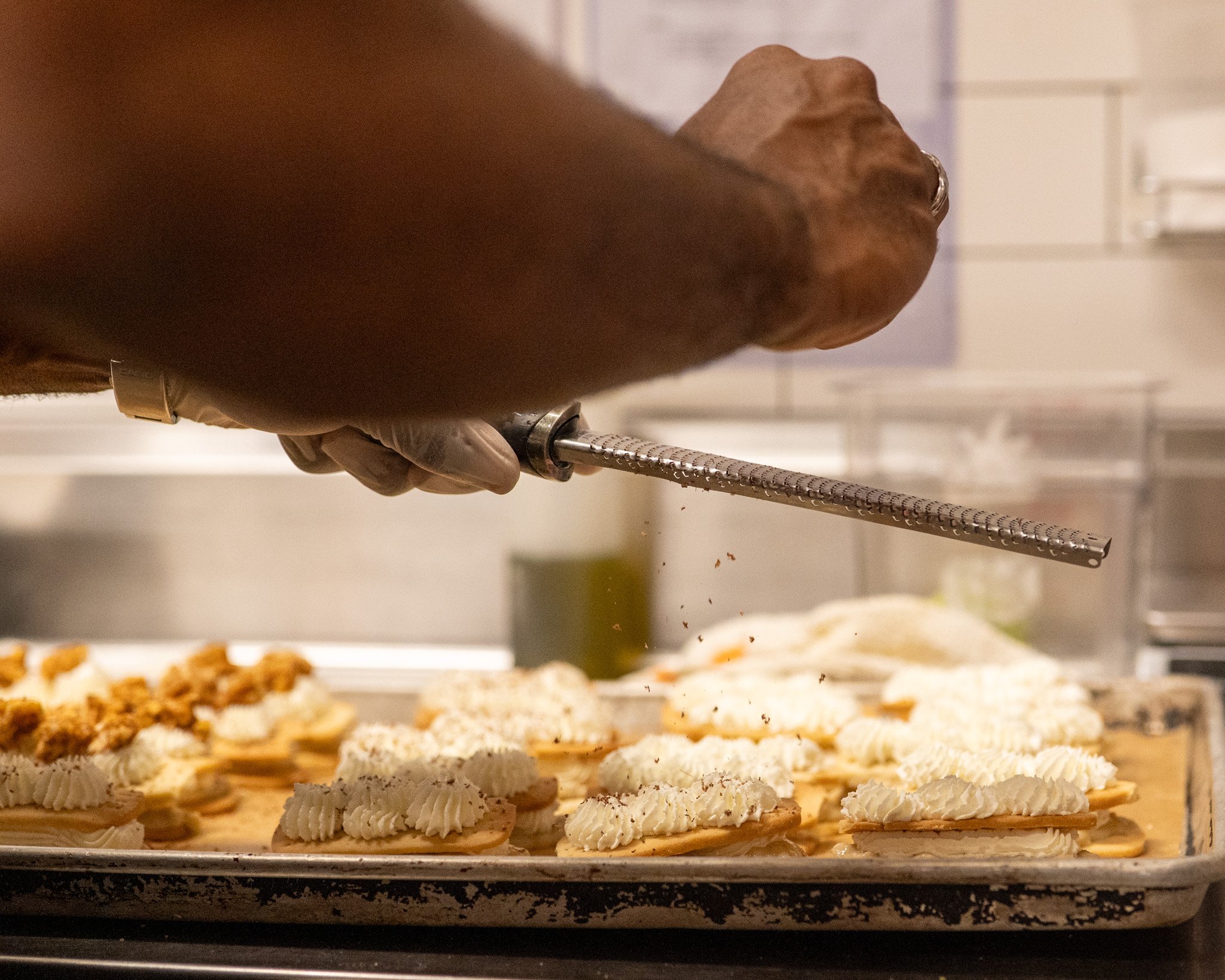 Hand holding a metal tool working on a layered flatbread or pastry with cream and toppings in a kitchen.