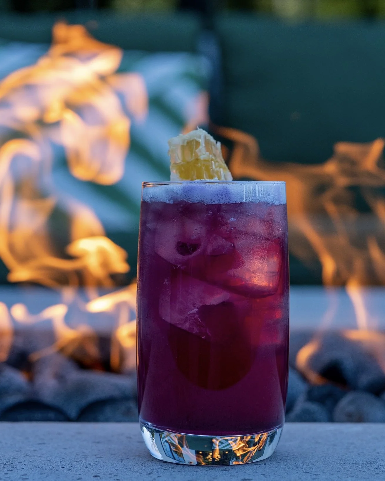 A tall glass filled with a purple beverage and ice cubes, garnished with a pineapple piece, with a fire pit in the background.