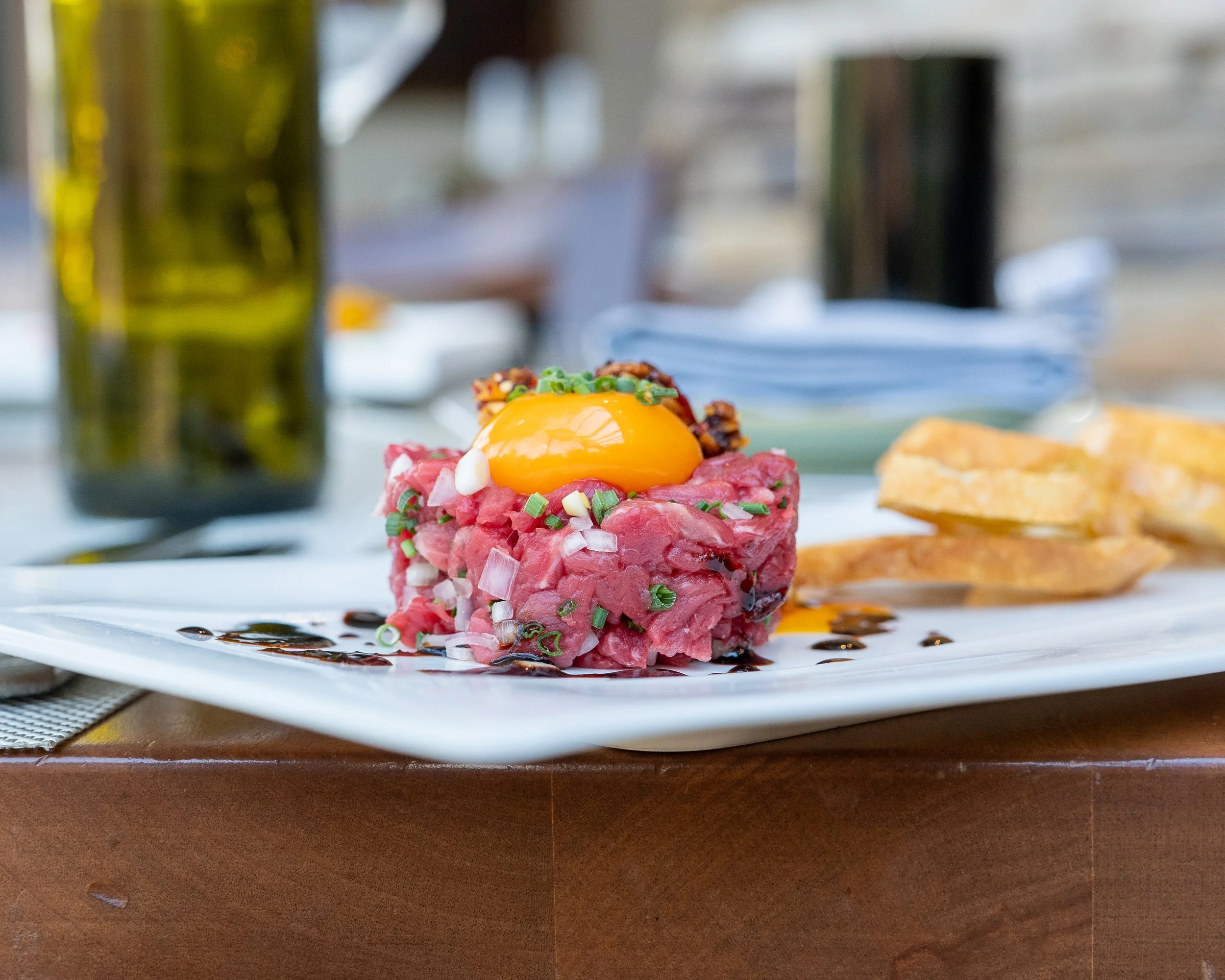 Close-up of beef tartare topped with raw egg yolk, chopped green onions, and crispy toppings on a white plate.