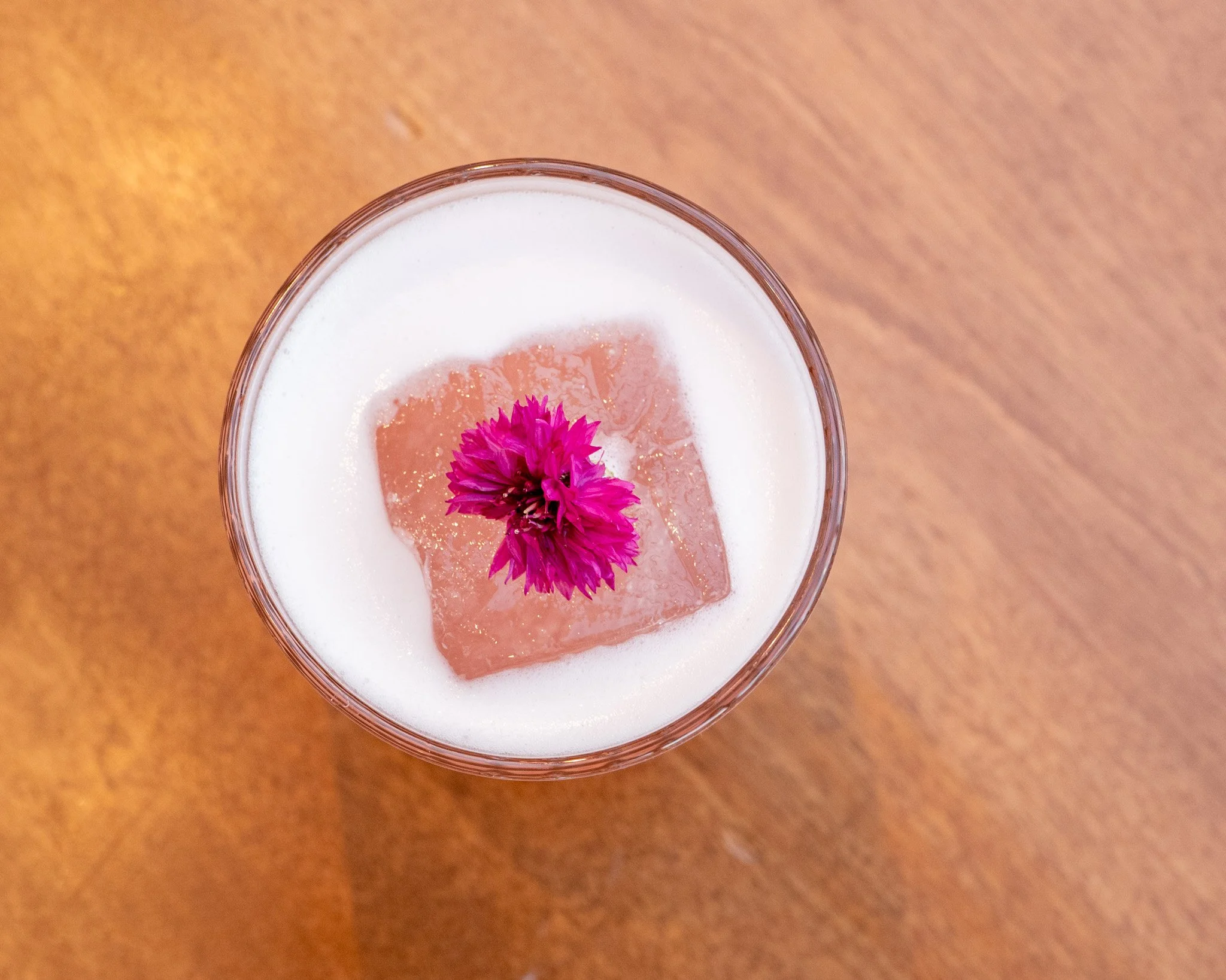A close-up of a glass of white beverage with a pink flower and a pink ice cube inside, placed on a wooden surface.