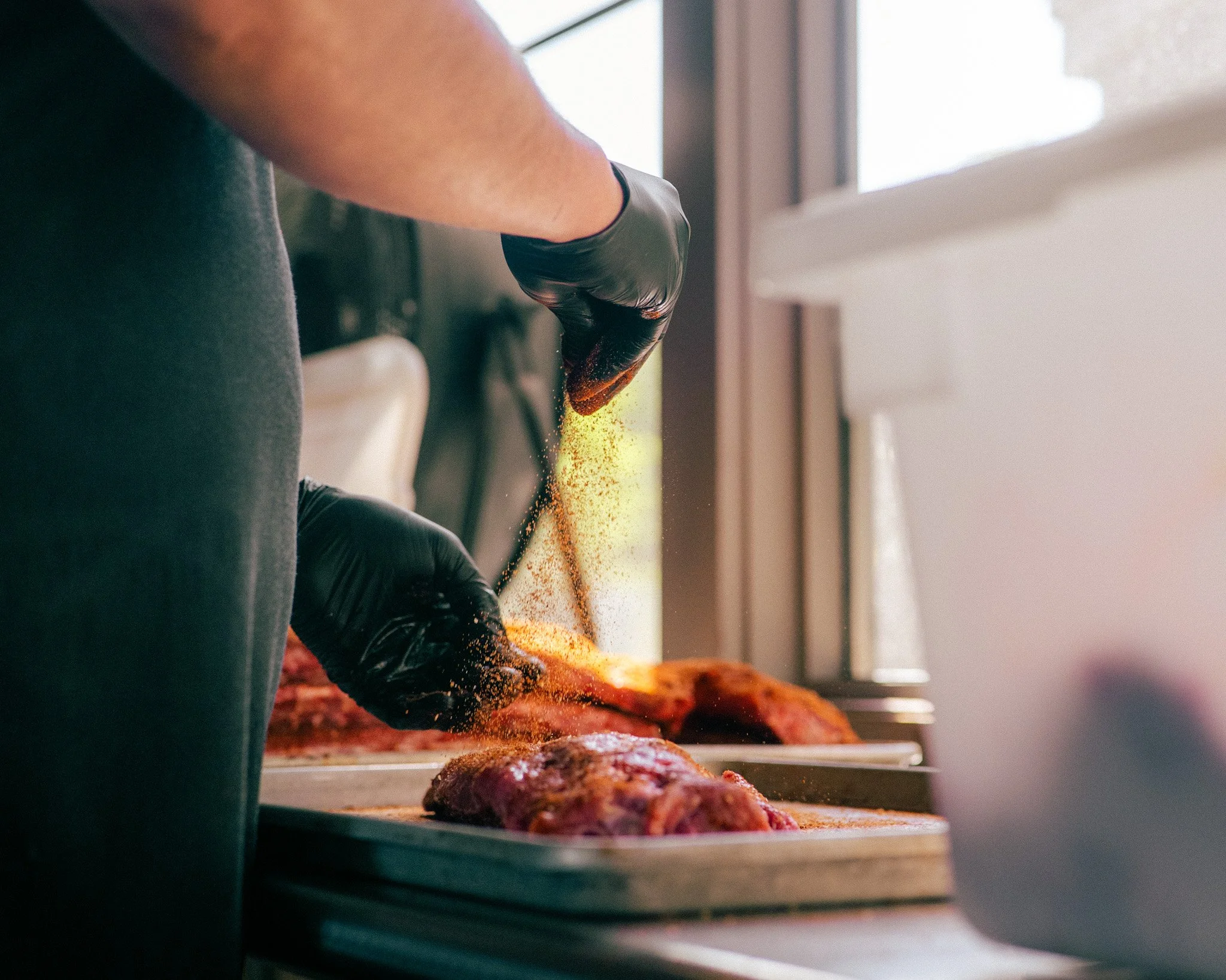 A person wearing black gloves is handling a large piece of smoked or roasted meat, possibly bacon or brisket, revealing its rich, dark color. The scene appears to be inside a kitchen or a barbecue area with natural light coming through a window.
