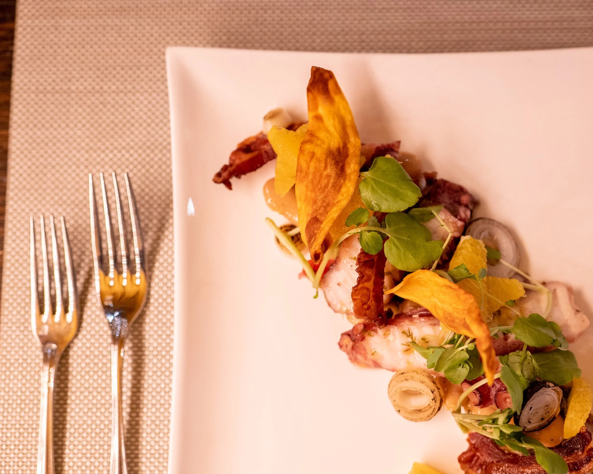 Close-up of a gourmet dish featuring grilled seafood, bacon, crispy fried plantain chips, and fresh greens on a white rectangular plate, with forks placed on a textured placemat nearby.
