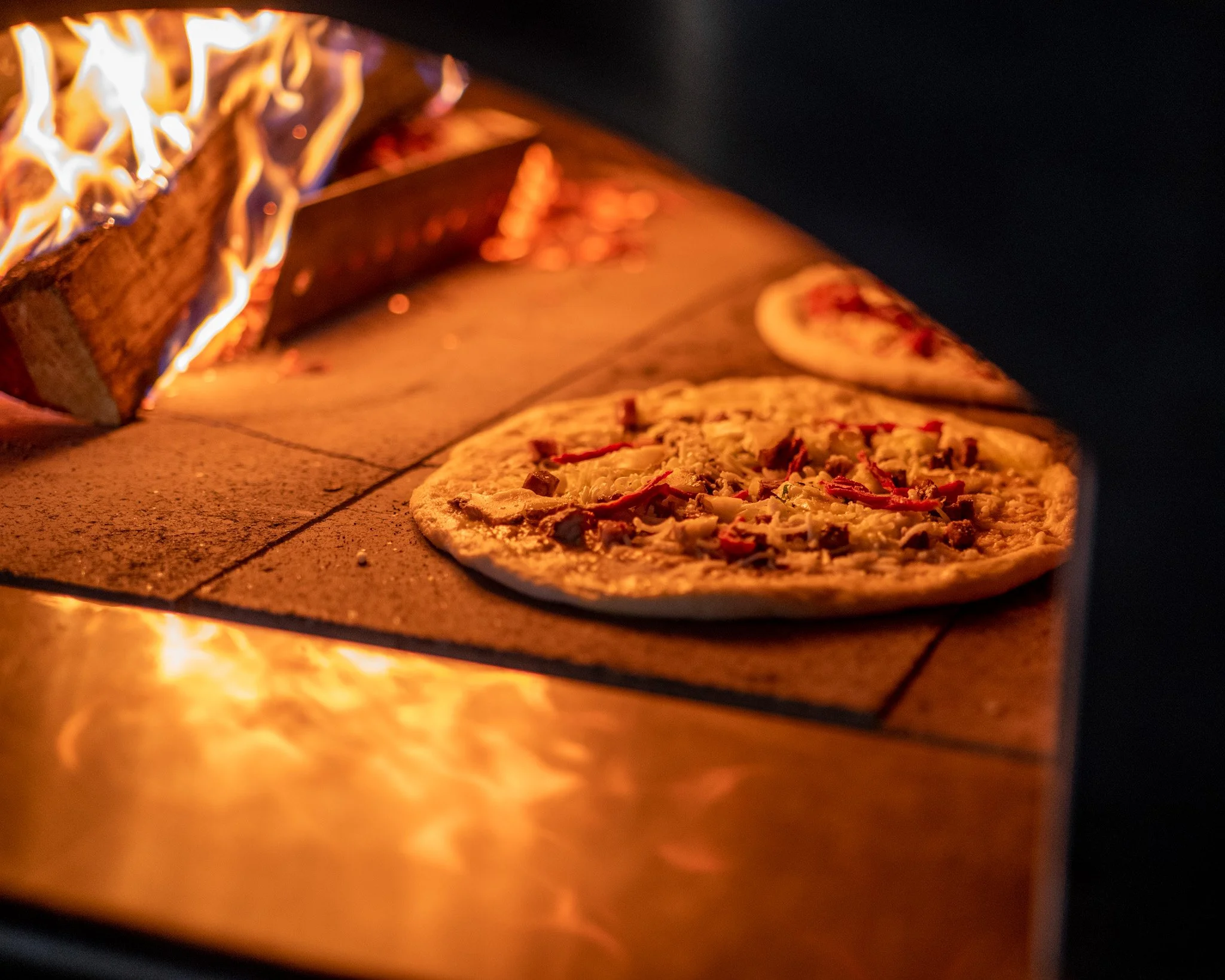 Pizza cooking in a wood-fired oven with flames visible in the background.