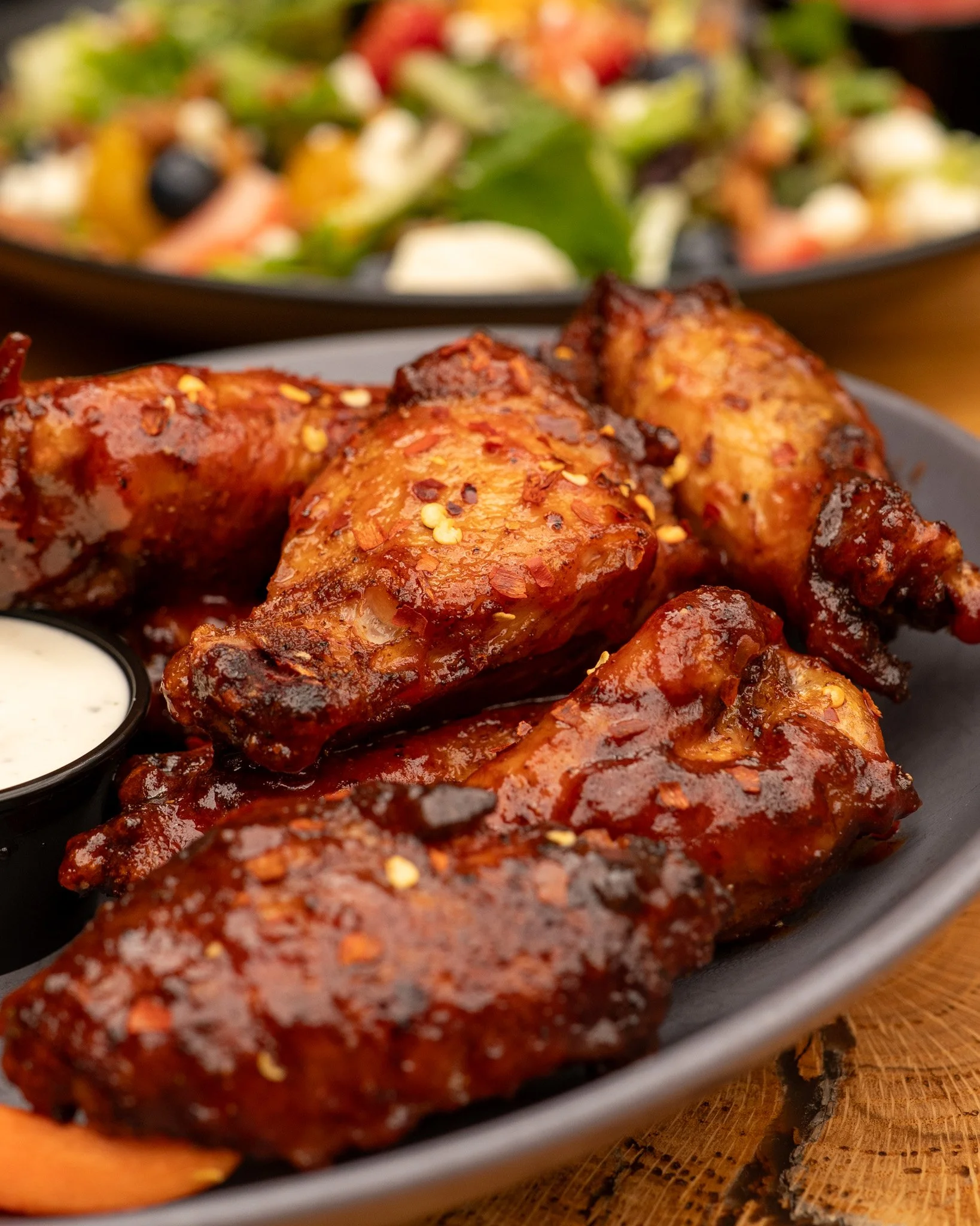 Close-up of glazed smoked chicken wings with chili flakes served on a black platter, with a side of creamy dipping sauce, and a colorful salad in the background.