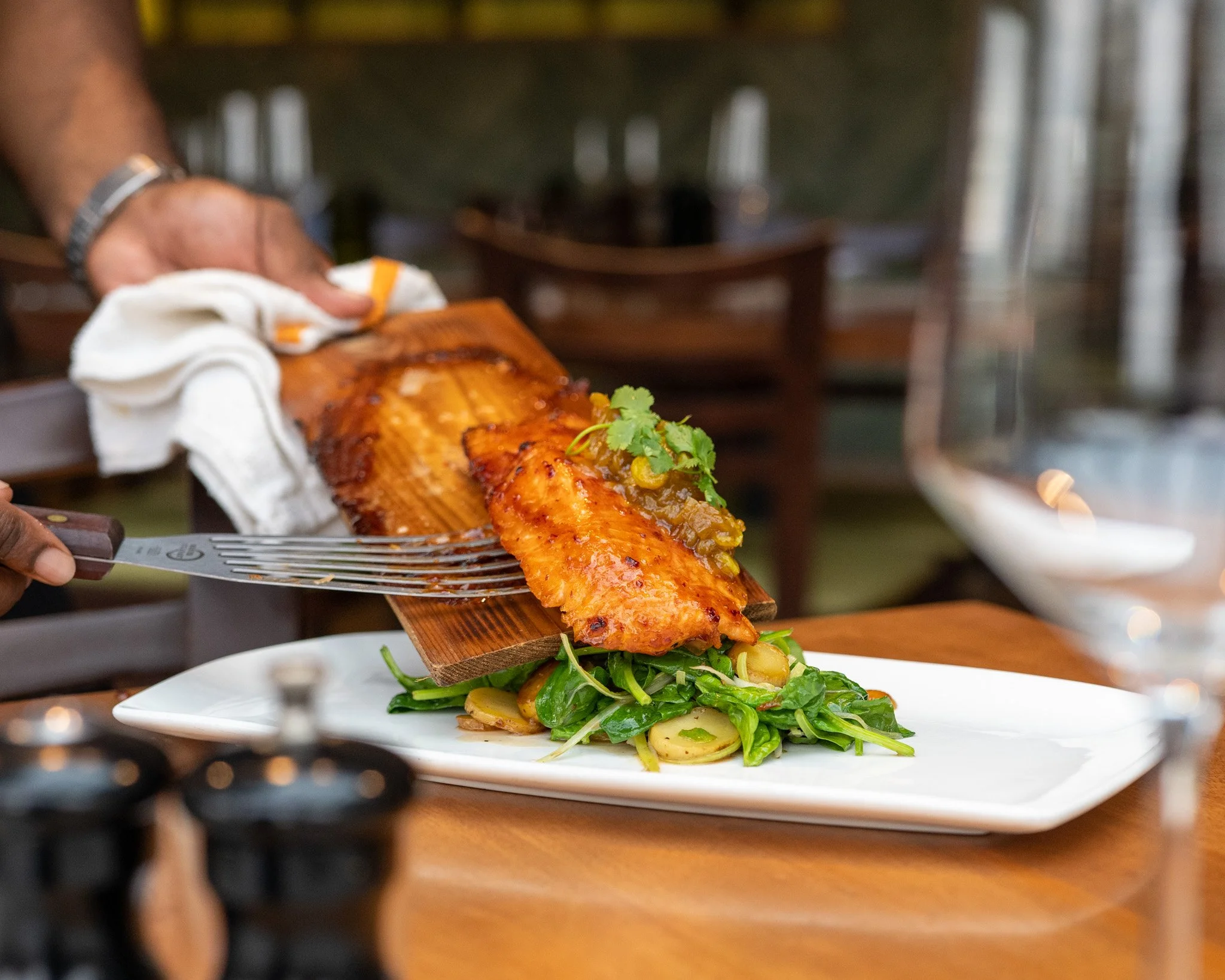 A person serving a grilled fish fillet with a garnish of cilantro onto a bed of greens on a white plate.