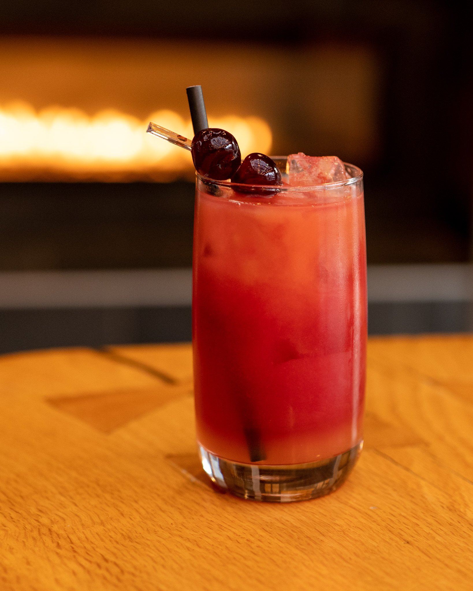 A tall glass filled with a red/pink beverage, ice cubes, and garnished with two dark cherries and a black straw, sitting on a wooden surface with a blurred warm-toned background.