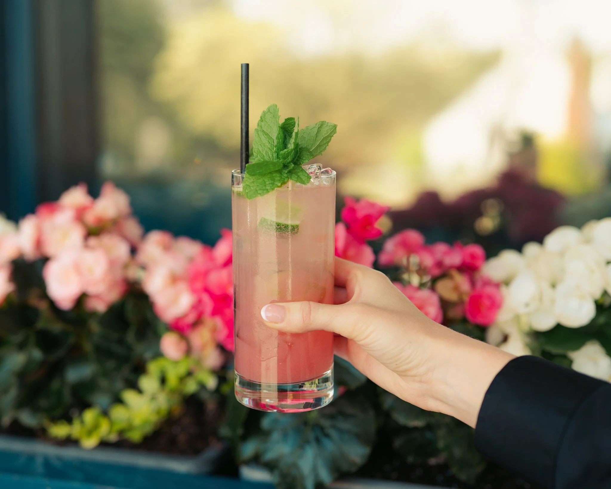 A person holding a tall glass of pink cocktail garnished with fresh mint leaves and a black straw, with pink and white flowers blurred in the background.