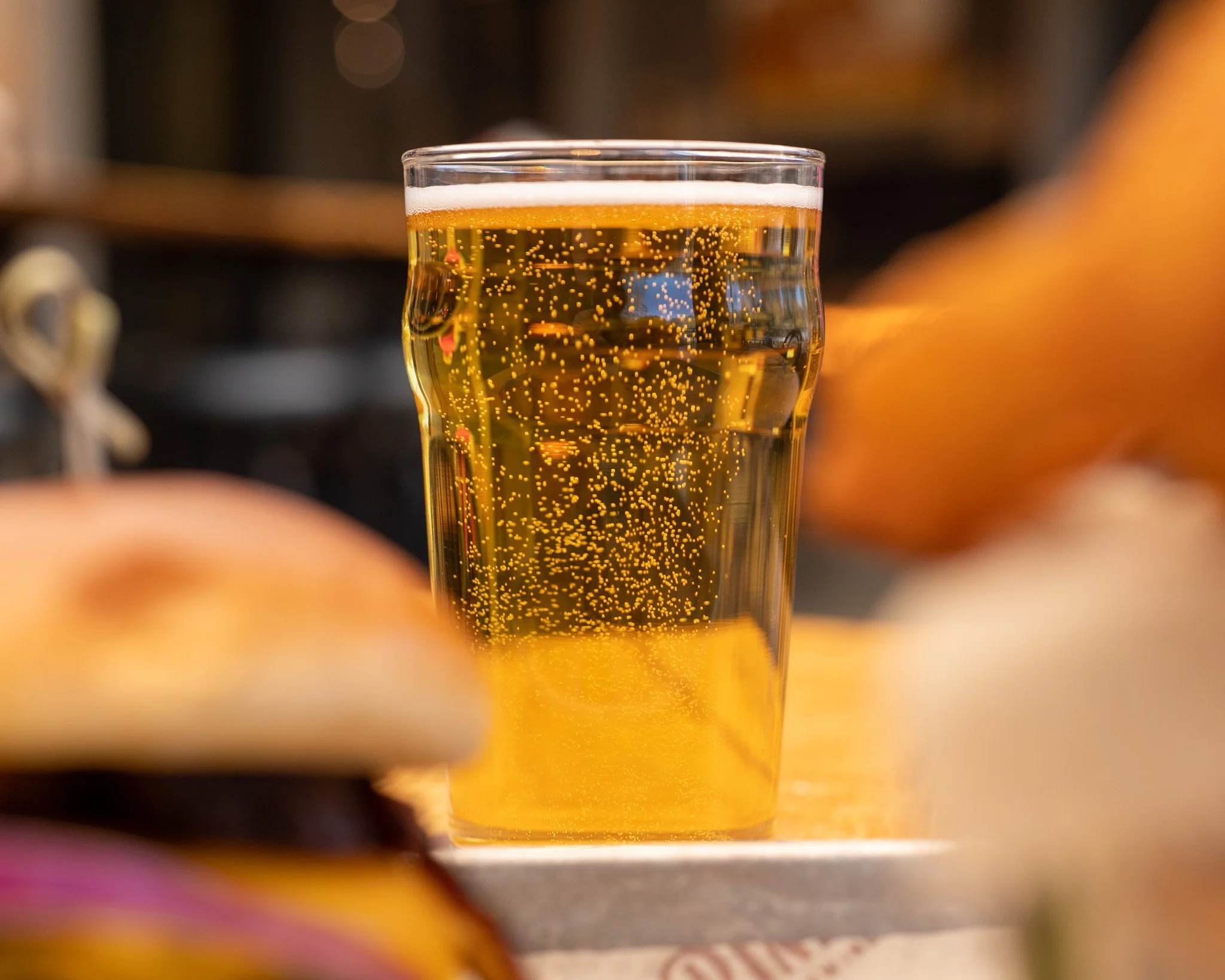 A clear glass filled with a bubbly, golden-colored beverage, likely beer, with a layer of foam on top and bubbles rising through the drink.