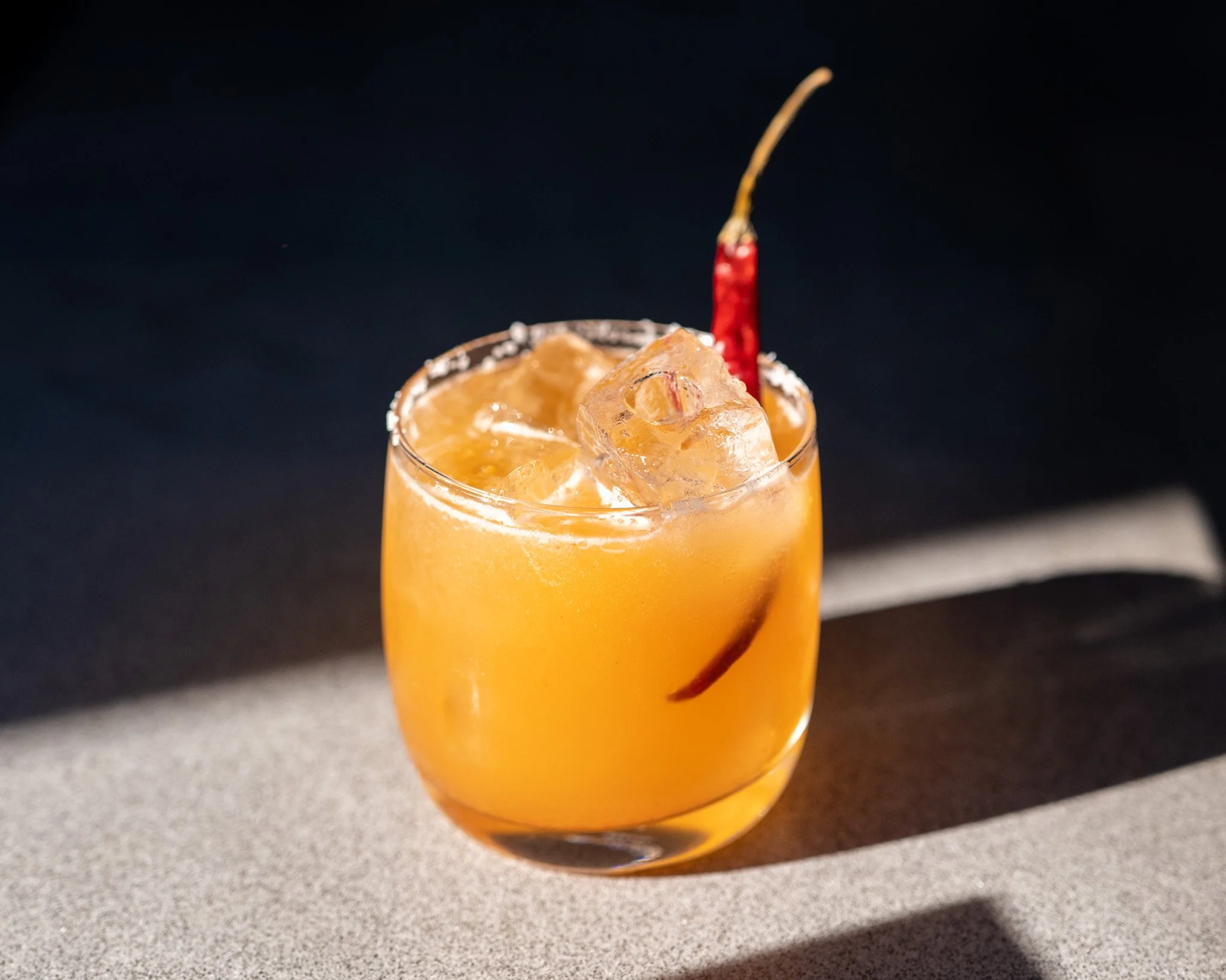 Close-up of a glass filled with orange-colored cocktail with ice cubes and a red chili pepper garnish against a dark background.