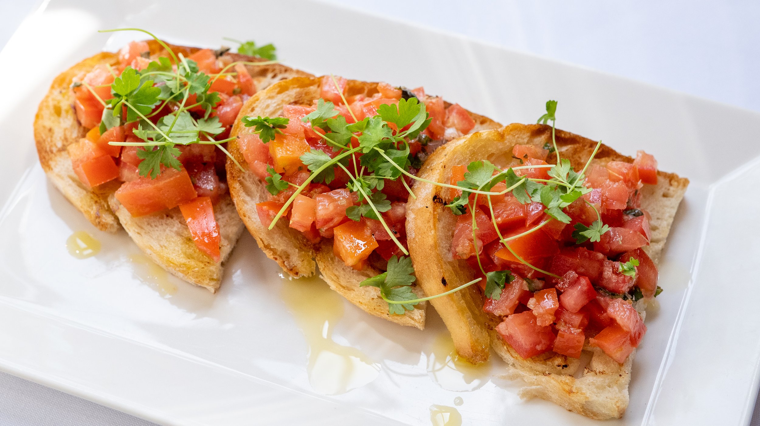 Three slices of toasted baguette topped with chopped tomatoes, herbs, and garnished with microgreens, drizzled with olive oil on a white rectangular plate.