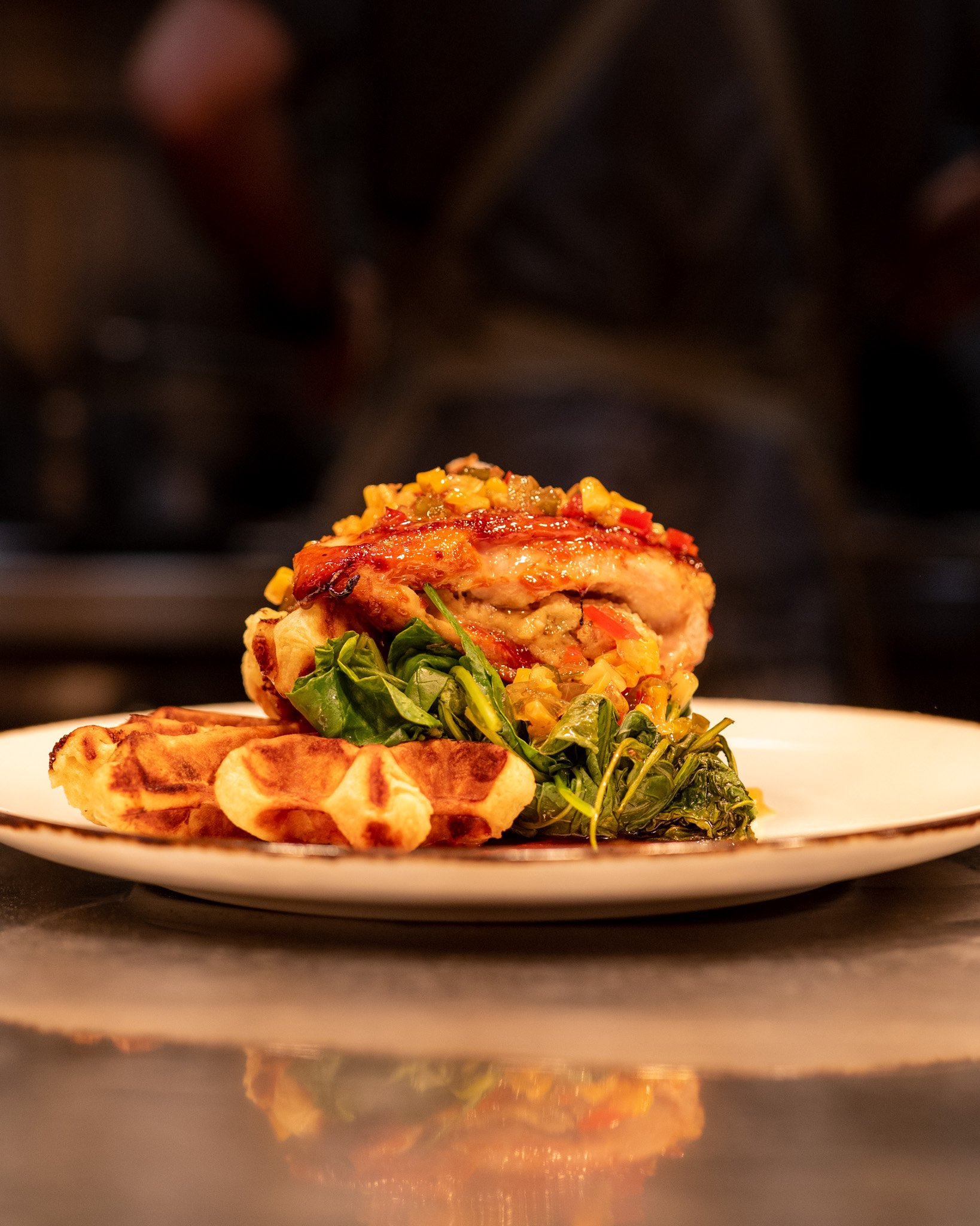 Close-up of a plated dish with a layered piece of grilled chicken topped with a vegetable salsa, accompanied by baked potato wedges and greens, with a dark background.