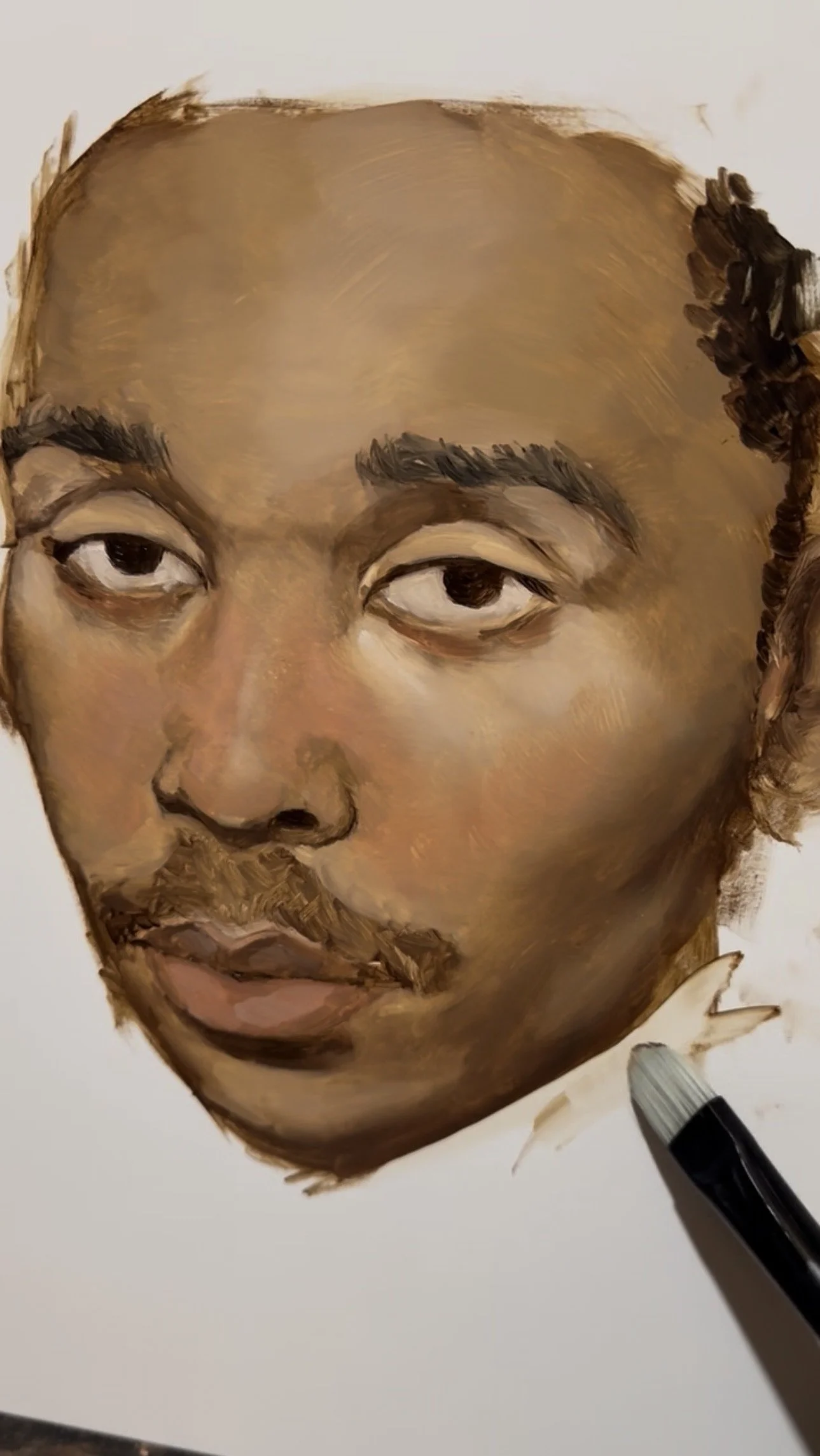 A close-up of a partially completed painted portrait of a person's face on a panel, with a paint brush visible at the bottom right corner.