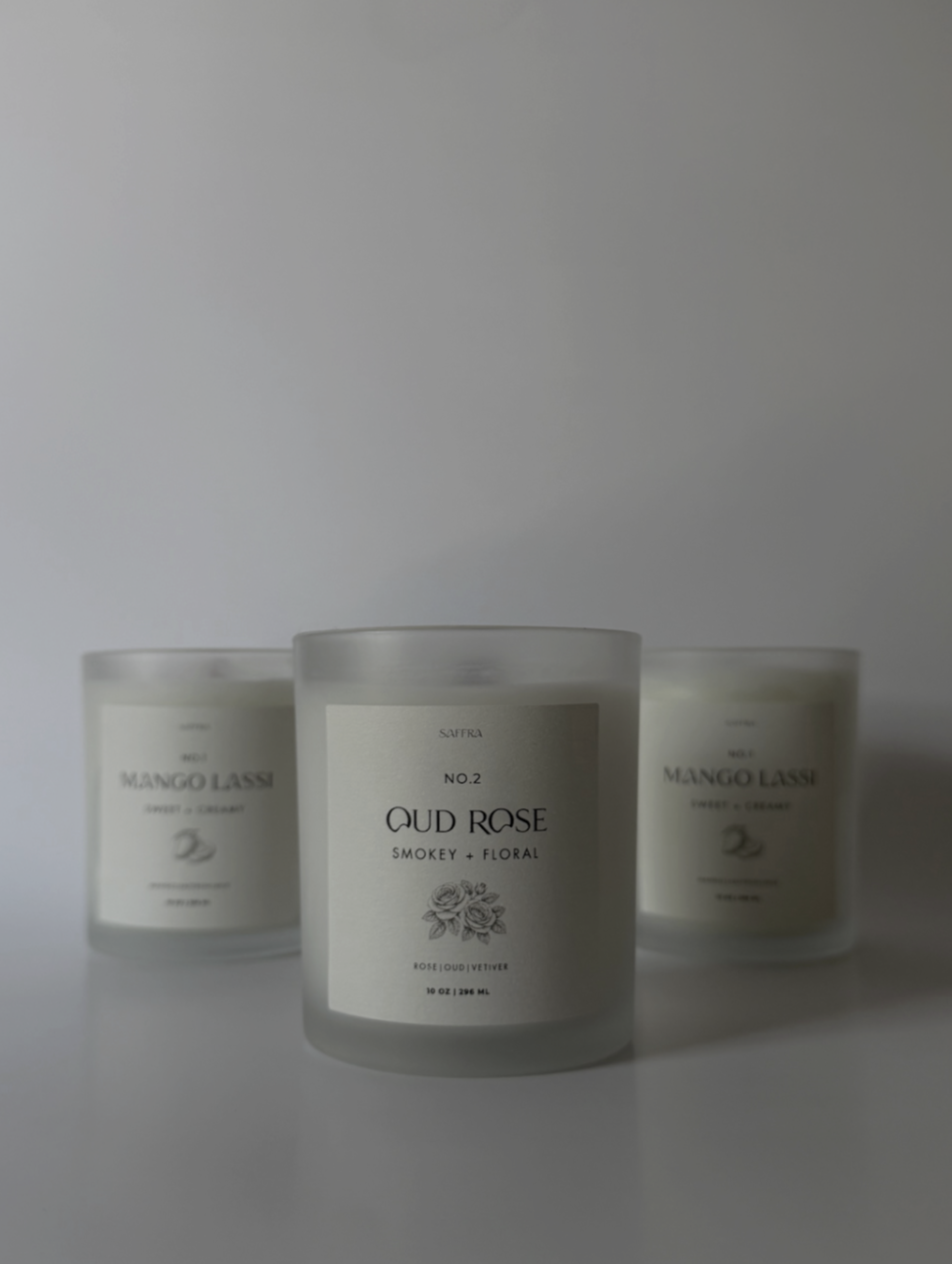 Three candles in frosted glass jars with white labels, the middle candle labeled 'Oud Rose' with a rose illustration, positioned on a white surface against a plain light gray background.