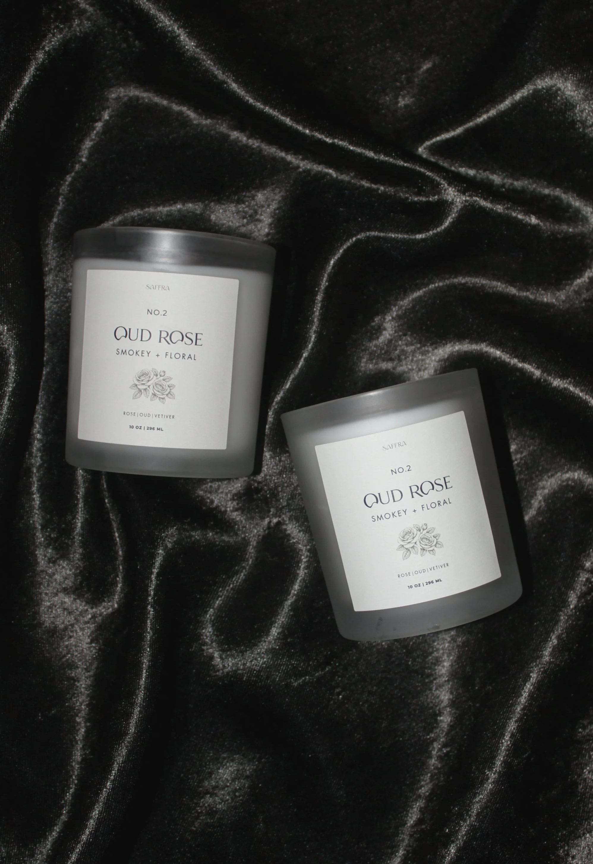 Two candles labeled 'Oud Rose Smokey + Floral' resting on black velvet fabric with shiny, textured surface.