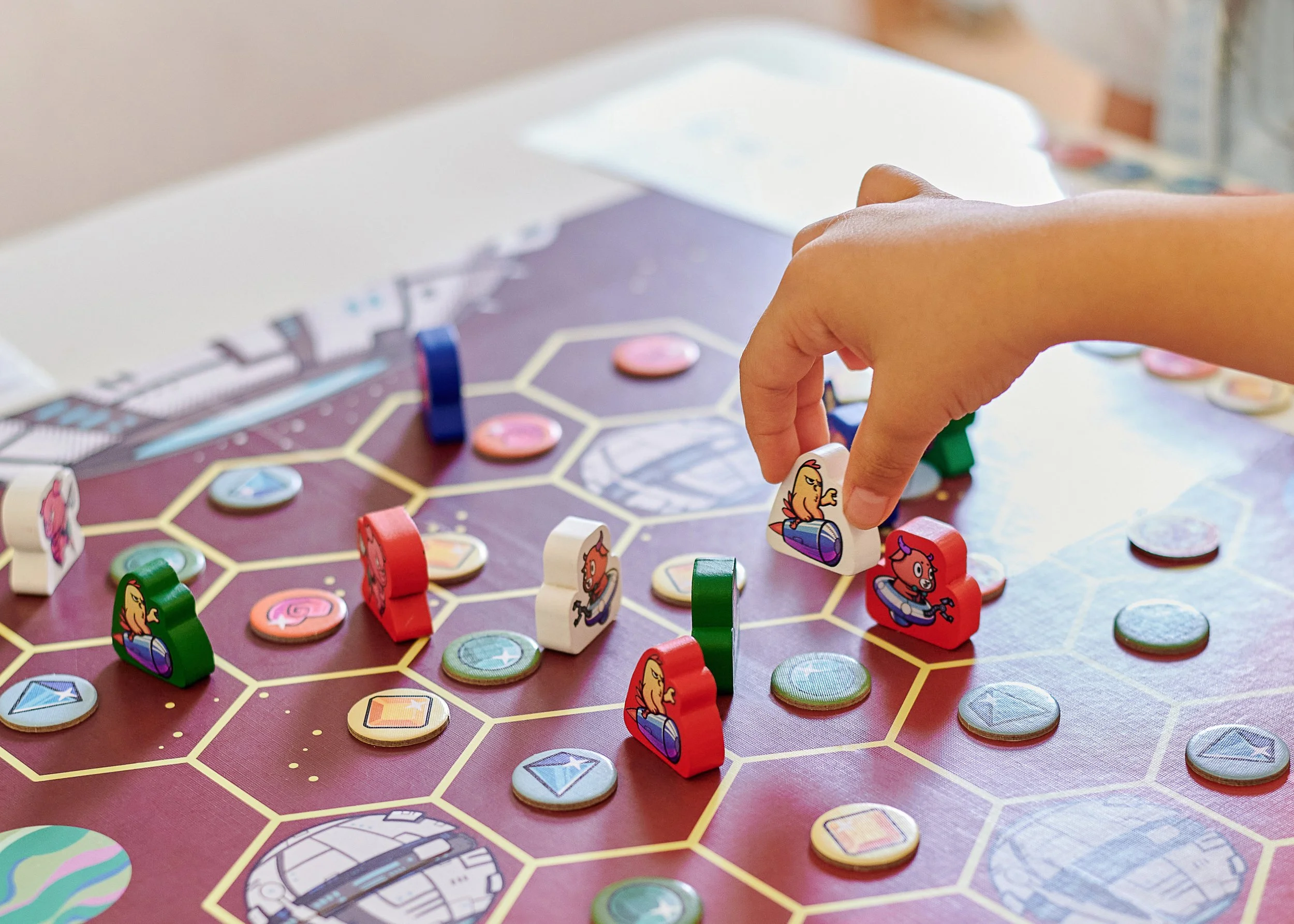 Close-up of a board game with hexagon tiles, game pieces, and cards, featuring a person's hand moving a game piece with a graphic of a chicken.