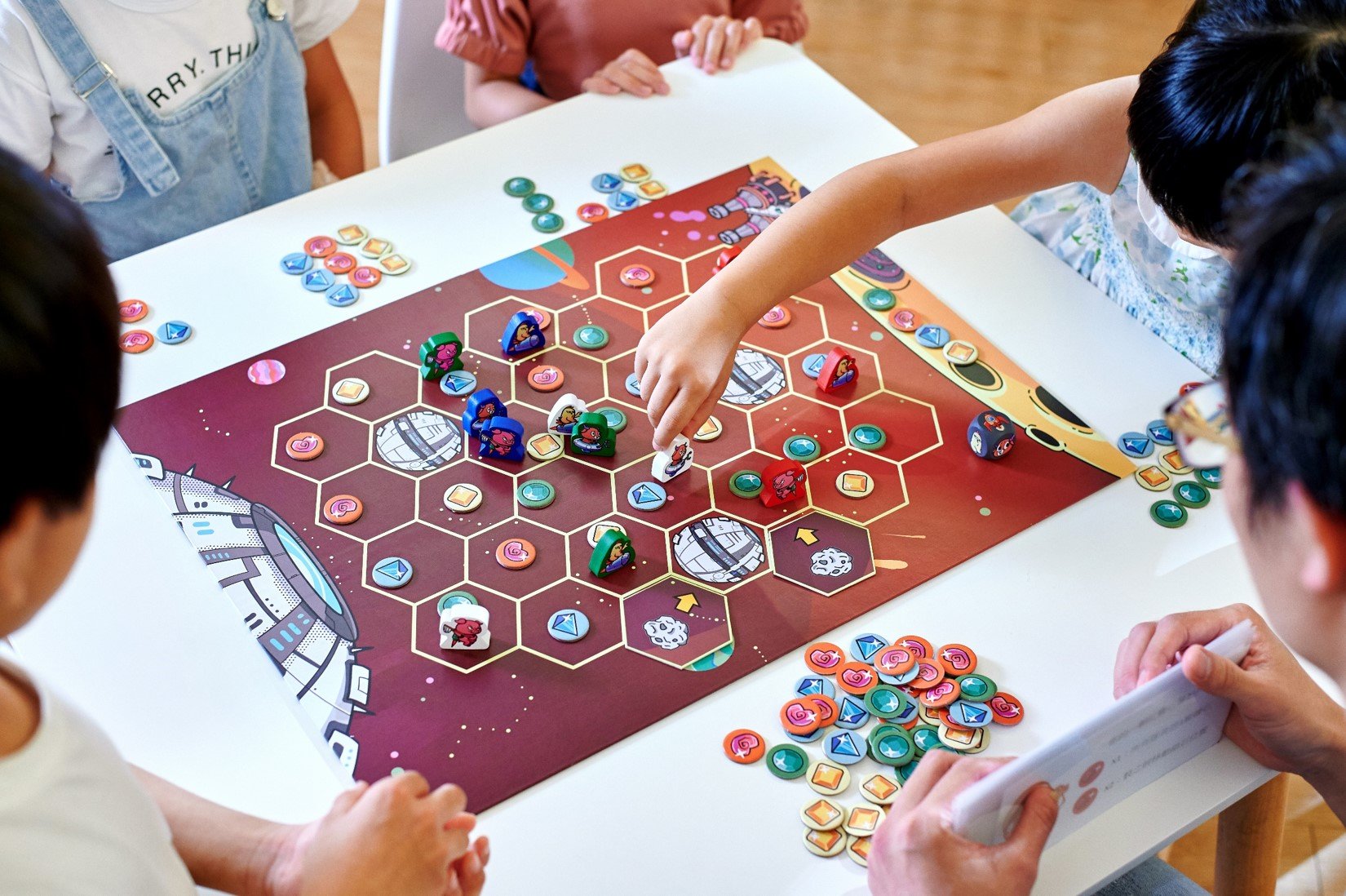 Children playing a space-themed board game with hexagonal tiles and colorful game pieces on a table.