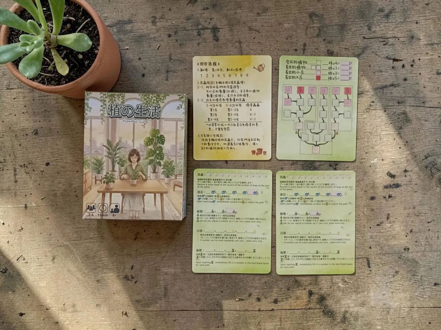 A tabletop view of a plant in a terracotta pot, a card game with Japanese instructions, and game components on a rustic wooden surface. The game box features an illustration of a woman sitting at a table surrounded by plants.