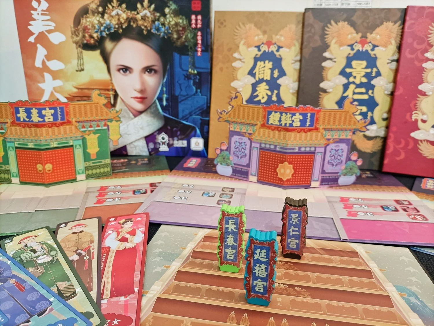 Photo of a board game set with colorful illustrated game pieces, cards, and game boards featuring traditional Chinese architecture and artwork, with a large box displaying a woman in traditional Chinese attire and headpiece.