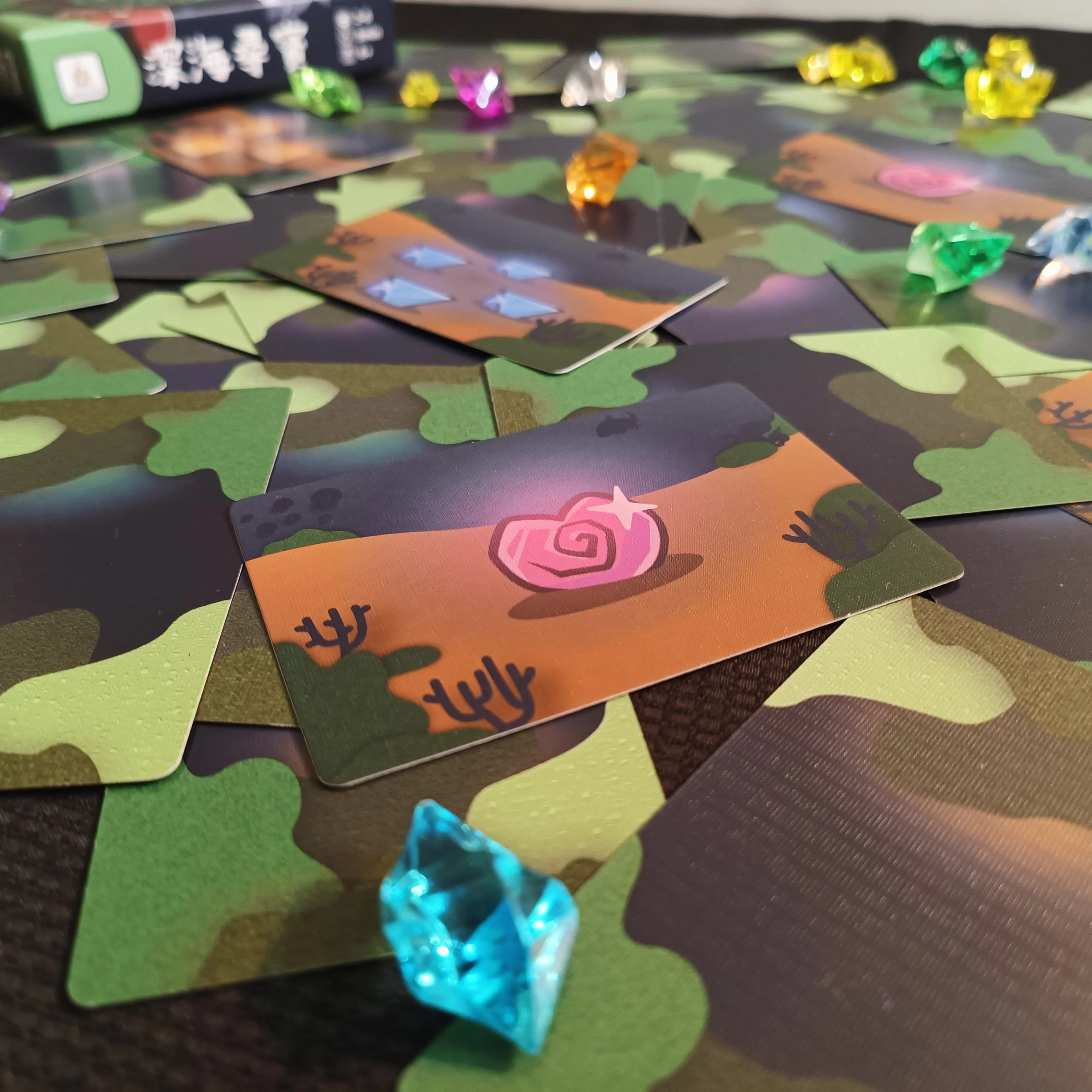 Close-up of scattered game pieces, including colorful dice and cards, on a camouflage-patterned surface.