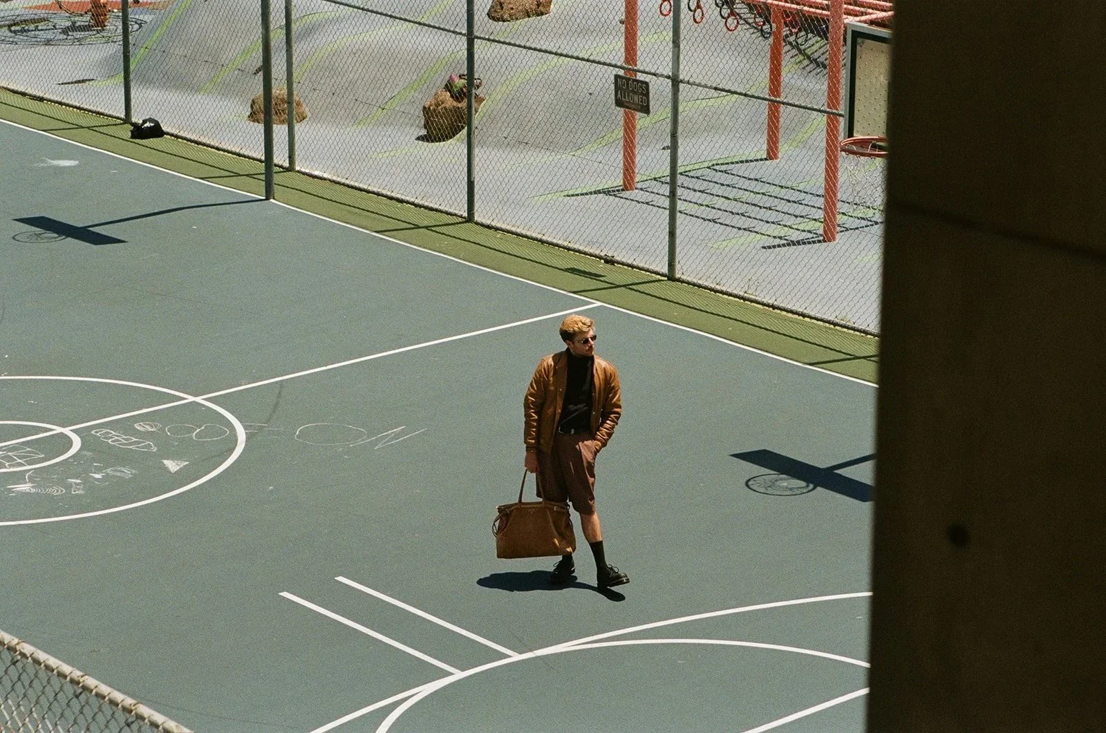 A man dressed in a brown jacket, black shirt, and shorts carrying a brown bag walking on an outdoor basketball court, with a chain-link fence and a sign that says 'No Dogs Allowed' behind him.