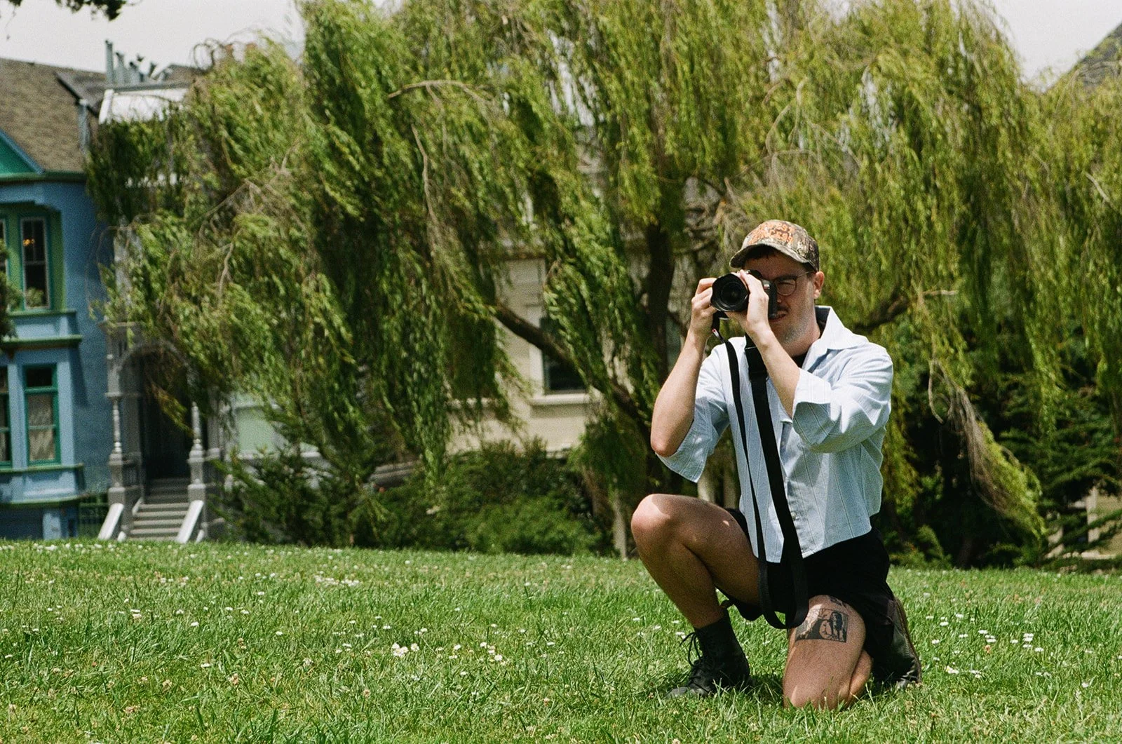 A man kneeling on a grassy lawn, taking a photograph with a camera. He is wearing glasses, a light-colored shirt, a baseball cap, and shorts with a tattoo visible on his thigh. In the background, there are trees and a blue house.