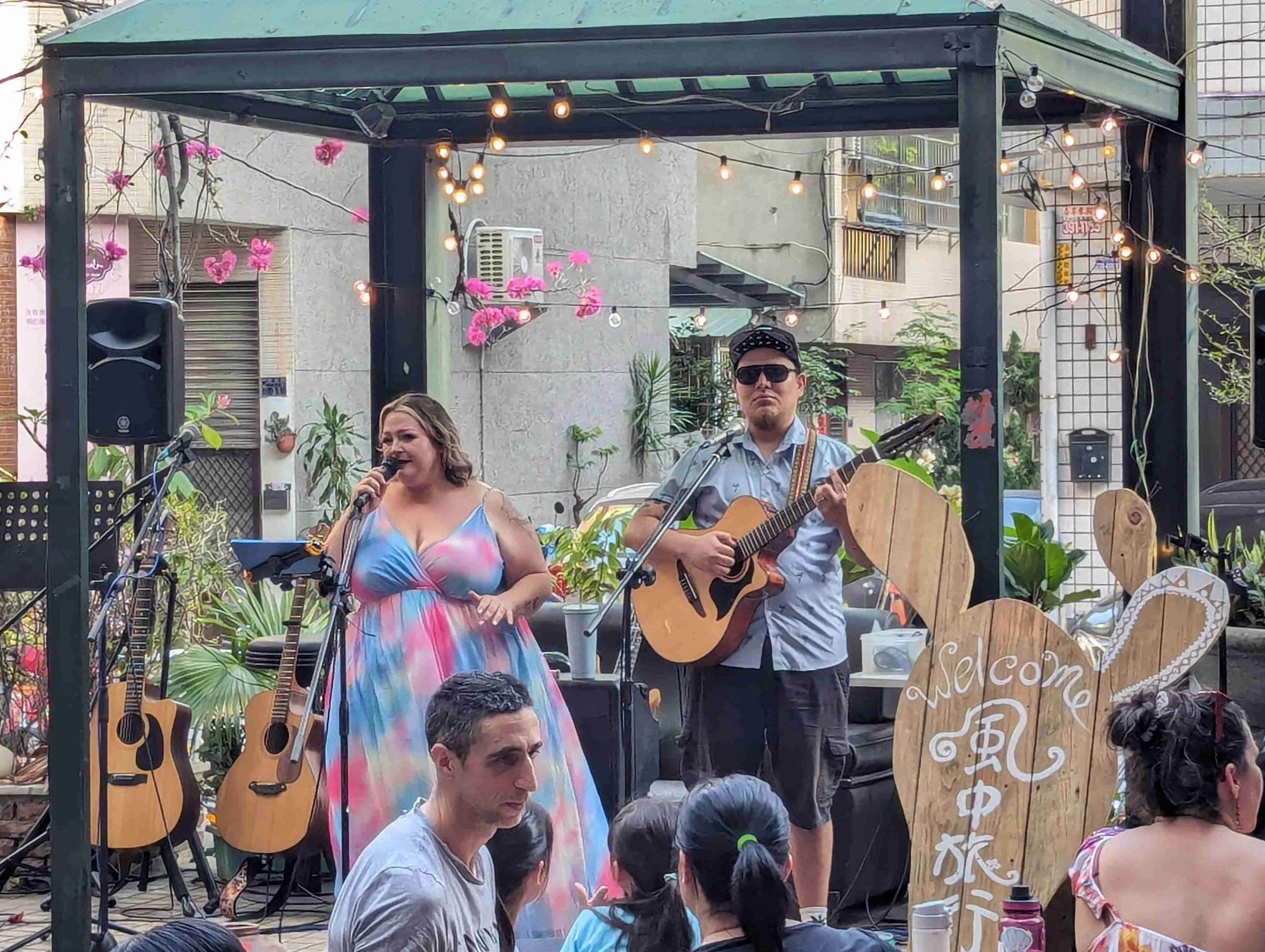 A small outdoor stage setup with two musicians performing: a woman singing into a microphone and a man playing an acoustic guitar. There are multiple acoustic guitars placed on stands on the stage. The stage is decorated with pink flowers and string 