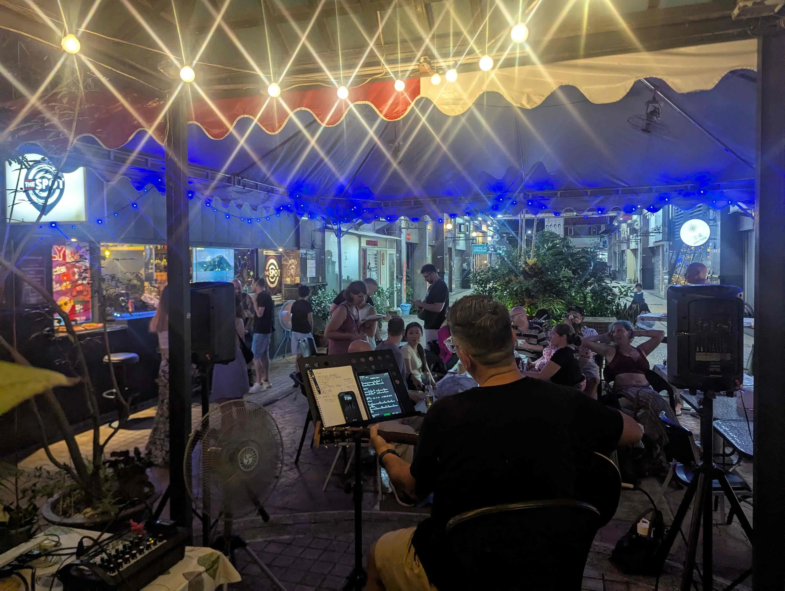 Night scene at an outdoor restaurant or bar with string lights overhead, bustling crowd, and a stage area with music equipment, people dining and socializing, illuminated signage and storefronts in the background.