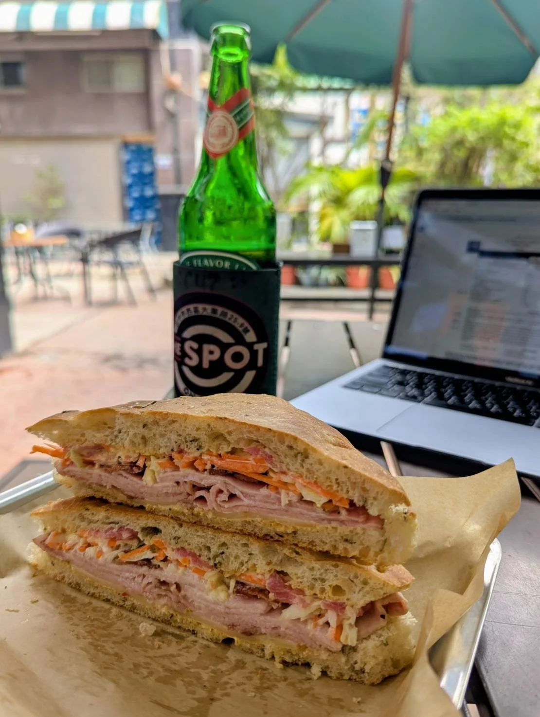 A club sandwich with layers of ham, turkey, cheese, and vegetables on toasted bread, placed on a tray. In the background, there is a green glass bottle of sparkling water in a black coaster with the logo 'The Spot', a laptop with a webpage open, and an outdoor seating area with tables, chairs, and an umbrella.