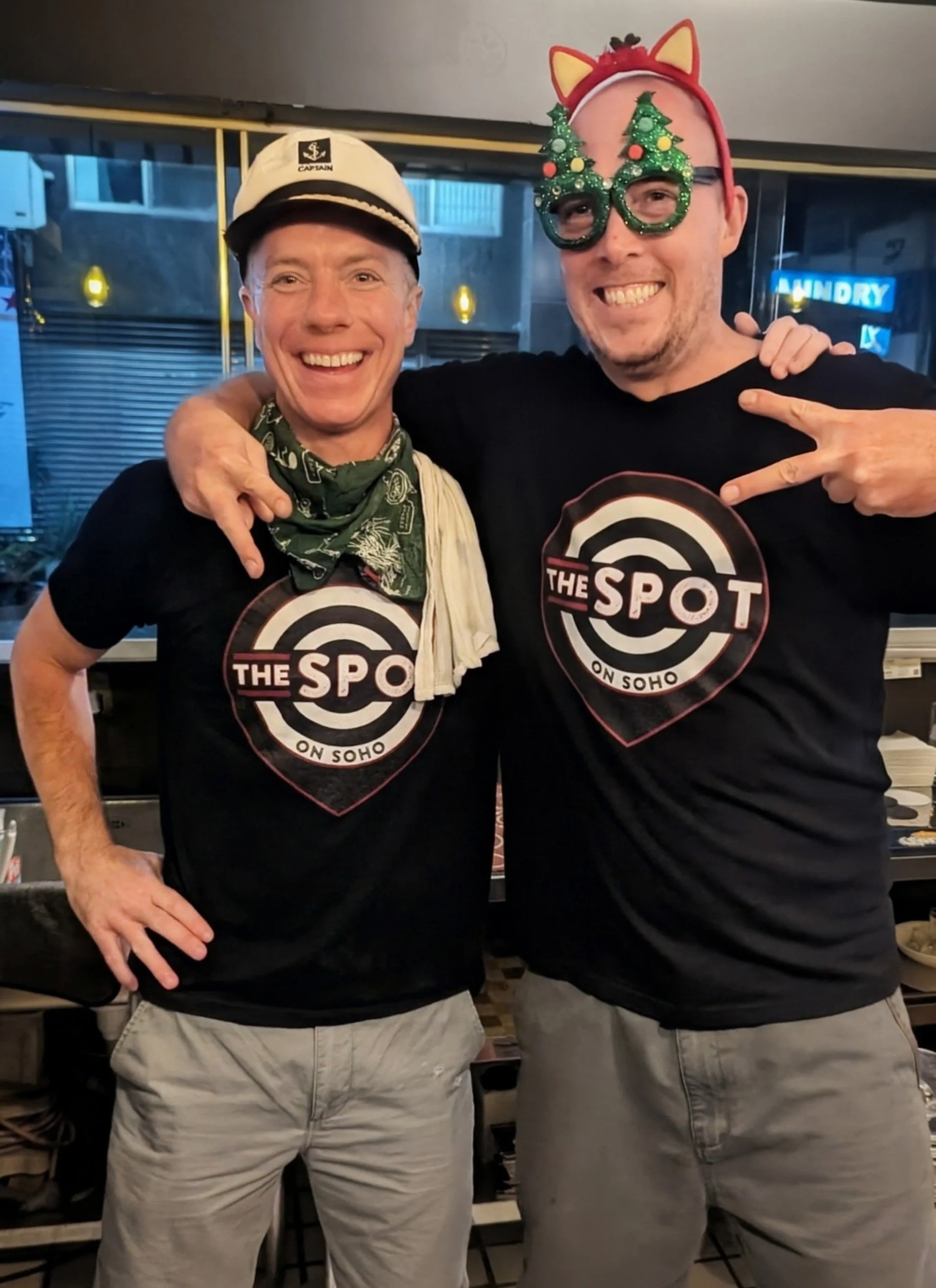 Two smiling men with their arms around each other, wearing black t-shirts with a circular logo reading 'The Spot on SoHo,' standing in a restaurant or bar. One man wears a captain's hat and a green bandana, the other has holiday glasses and a headband with festive Christmas trees. In the background, there is a sign that says 'LAUNDRY'.