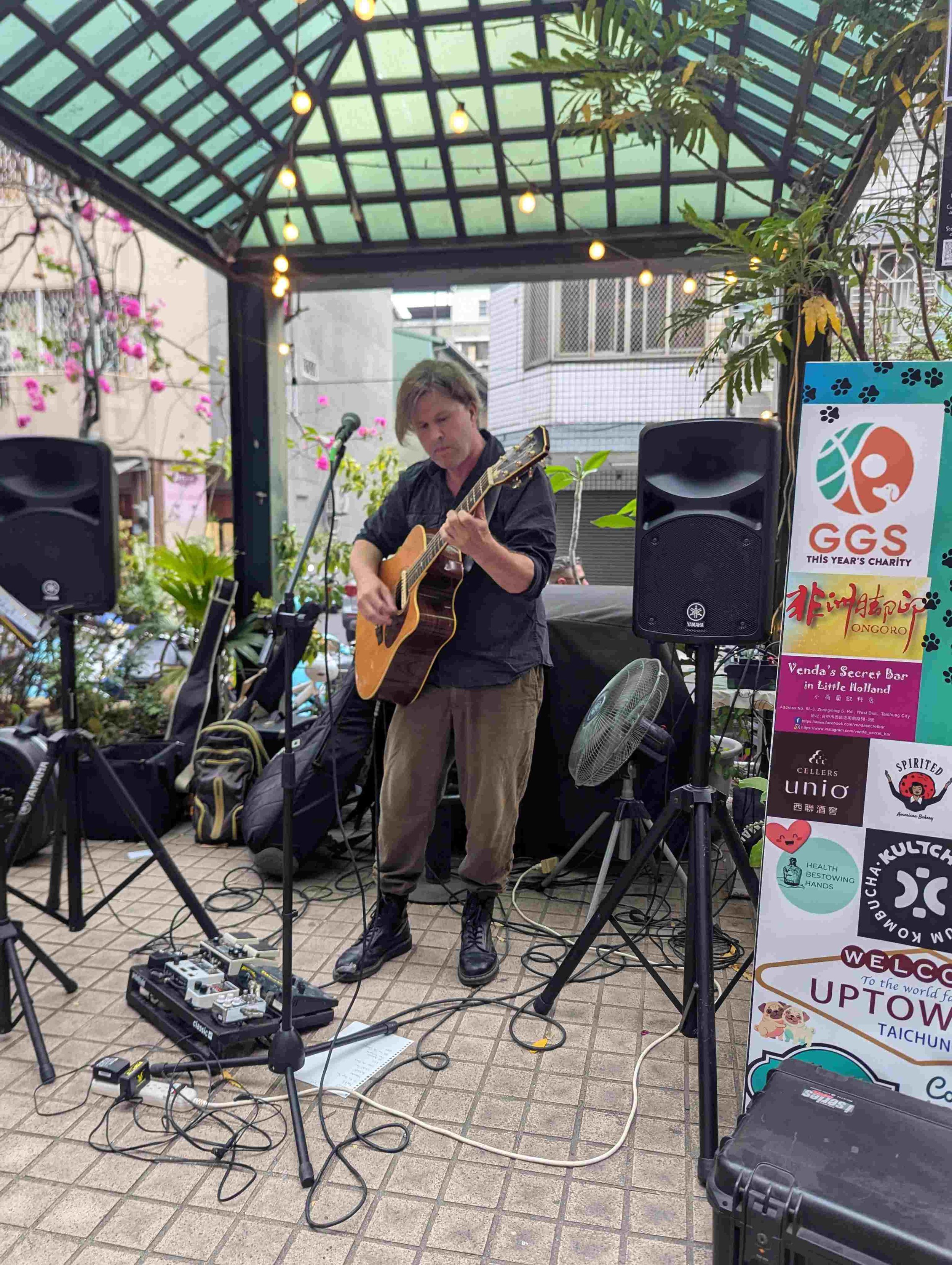A man playing an acoustic guitar in an outdoor setting under a green-tinted glass roof, with string lights overhead and a colorful charity sign nearby.