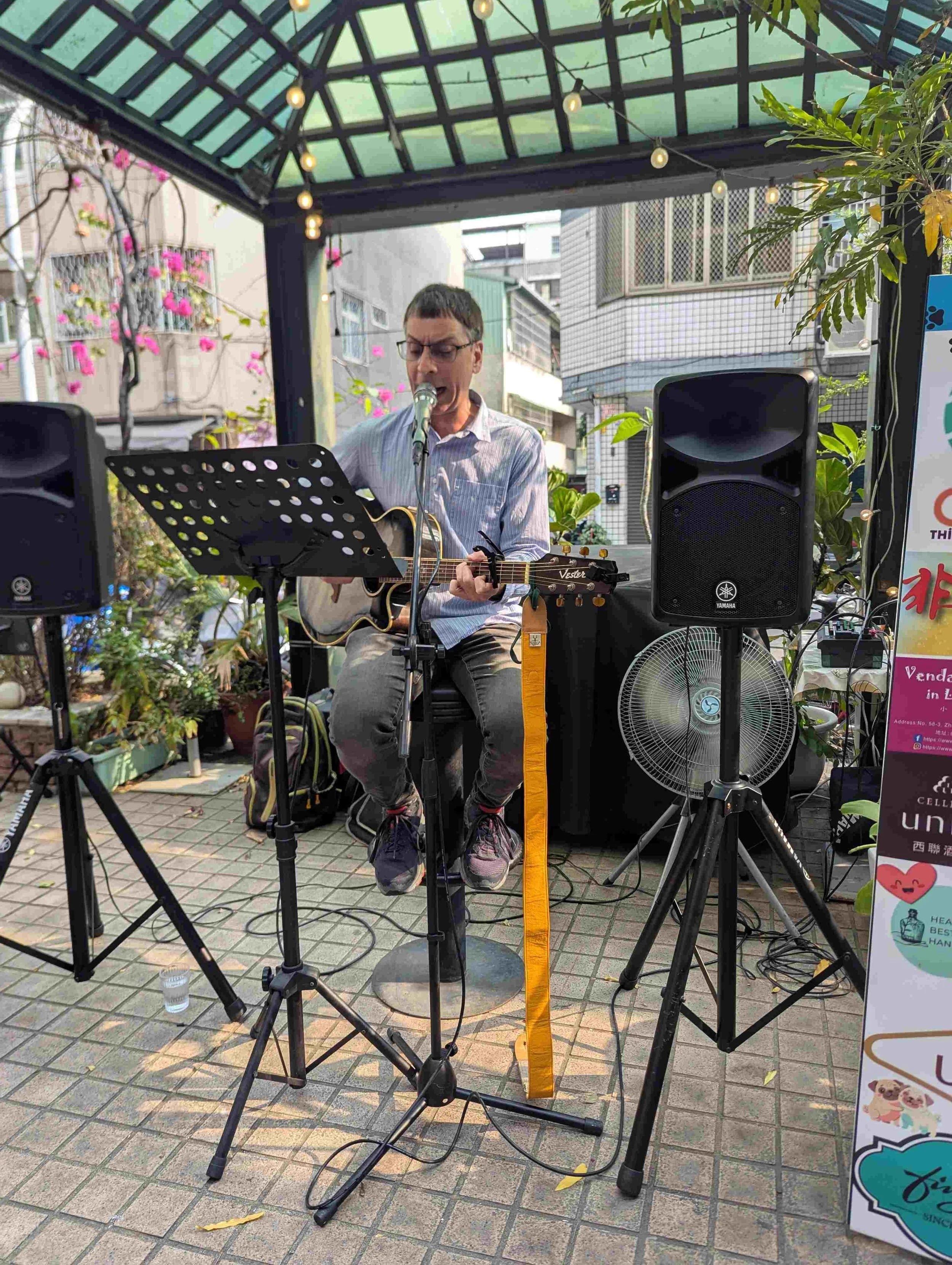 A man with glasses and a striped shirt is performing a song and playing an acoustic guitar under a canopy with string lights. There are two large speakers on either side, and a music stand in front of him. The background includes buildings, flowering