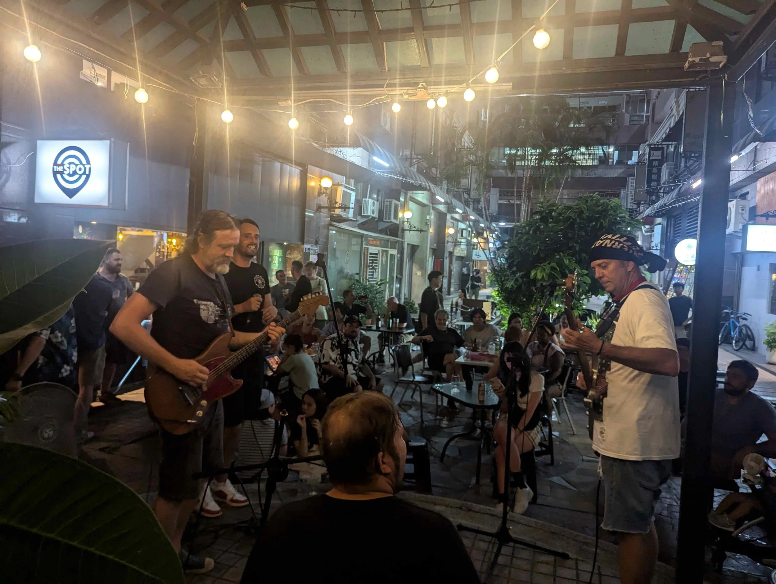 A nighttime outdoor dining scene with a live band playing music. The band consists of a guitarist and a vocalist, with a crowd of people enjoying the performance. String lights hang overhead, creating a warm ambiance. The setting appears to be a stre