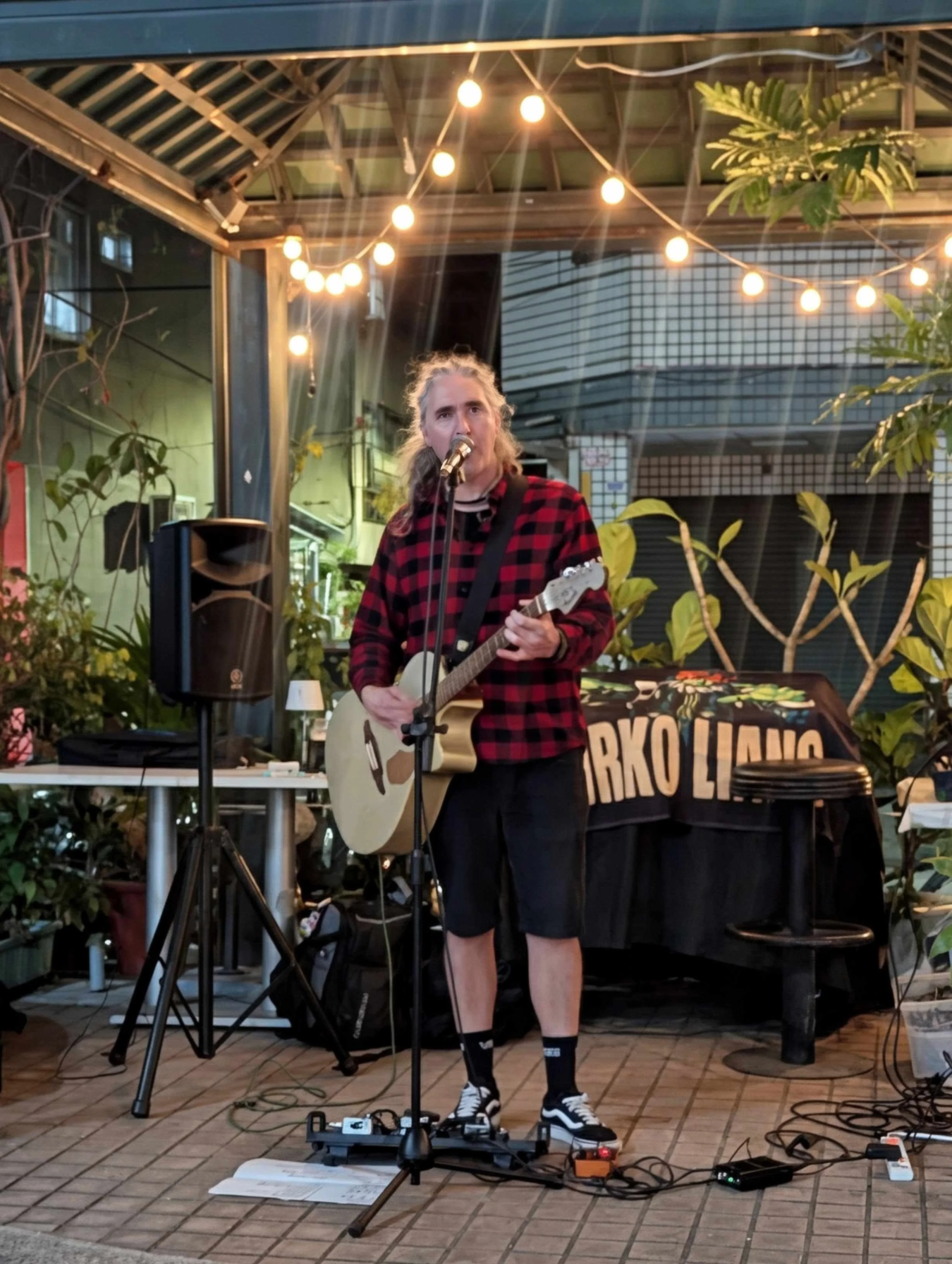 A person with long hair, wearing a red and black plaid shirt, shorts, and sneakers, is performing with a guitar and singing into a microphone in an outdoor setting decorated with string lights, plants, and a banner that reads "BARKO LUNG."