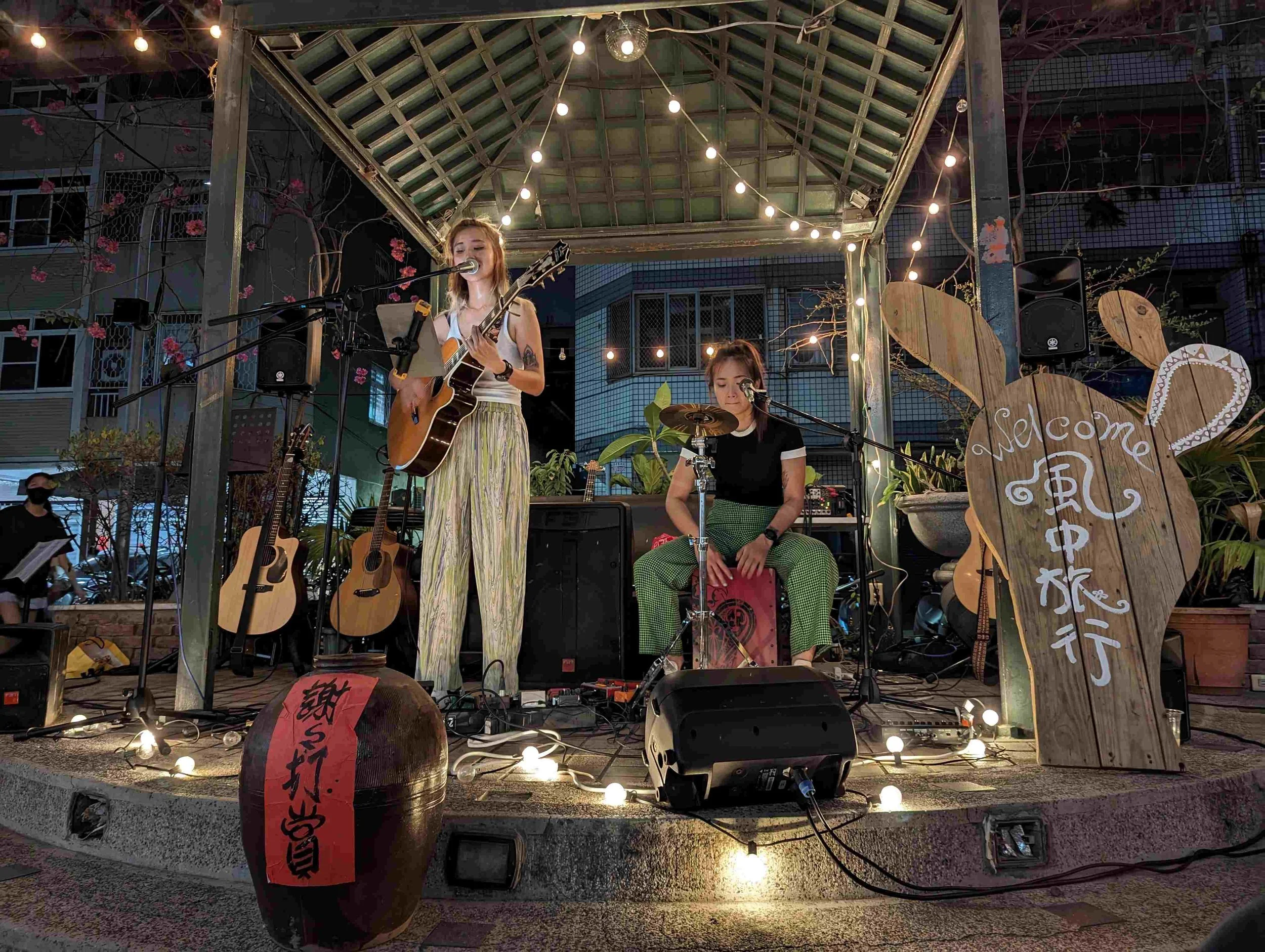A woman singing and playing guitar on an outdoor stage at night with string lights overhead, a man playing percussion, and musical instruments including guitars around them, surrounded by decorative plants and a wooden sign with Chinese characters.