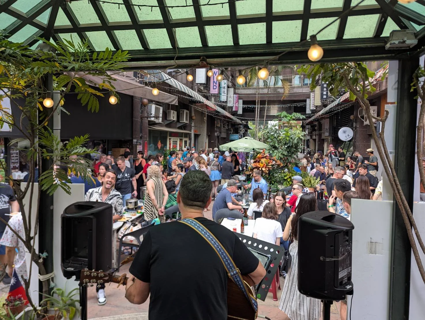 Crowd gathered in an outdoor restaurant or cafe, with a musician playing guitar in the foreground, surrounded by people enjoying food and drinks, and string lights hanging overhead.