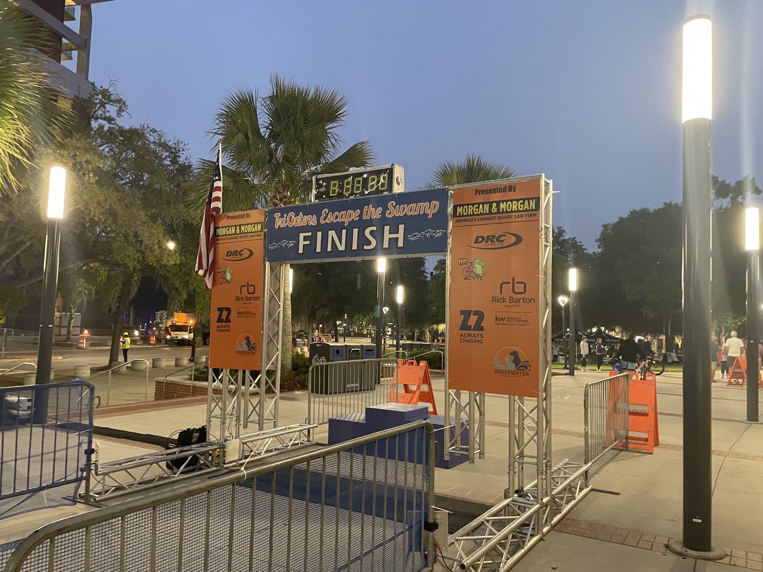 An outdoor scene at dusk showing a finish line banner for a race event, with palm trees and street lights illuminating the area and some people in the background.