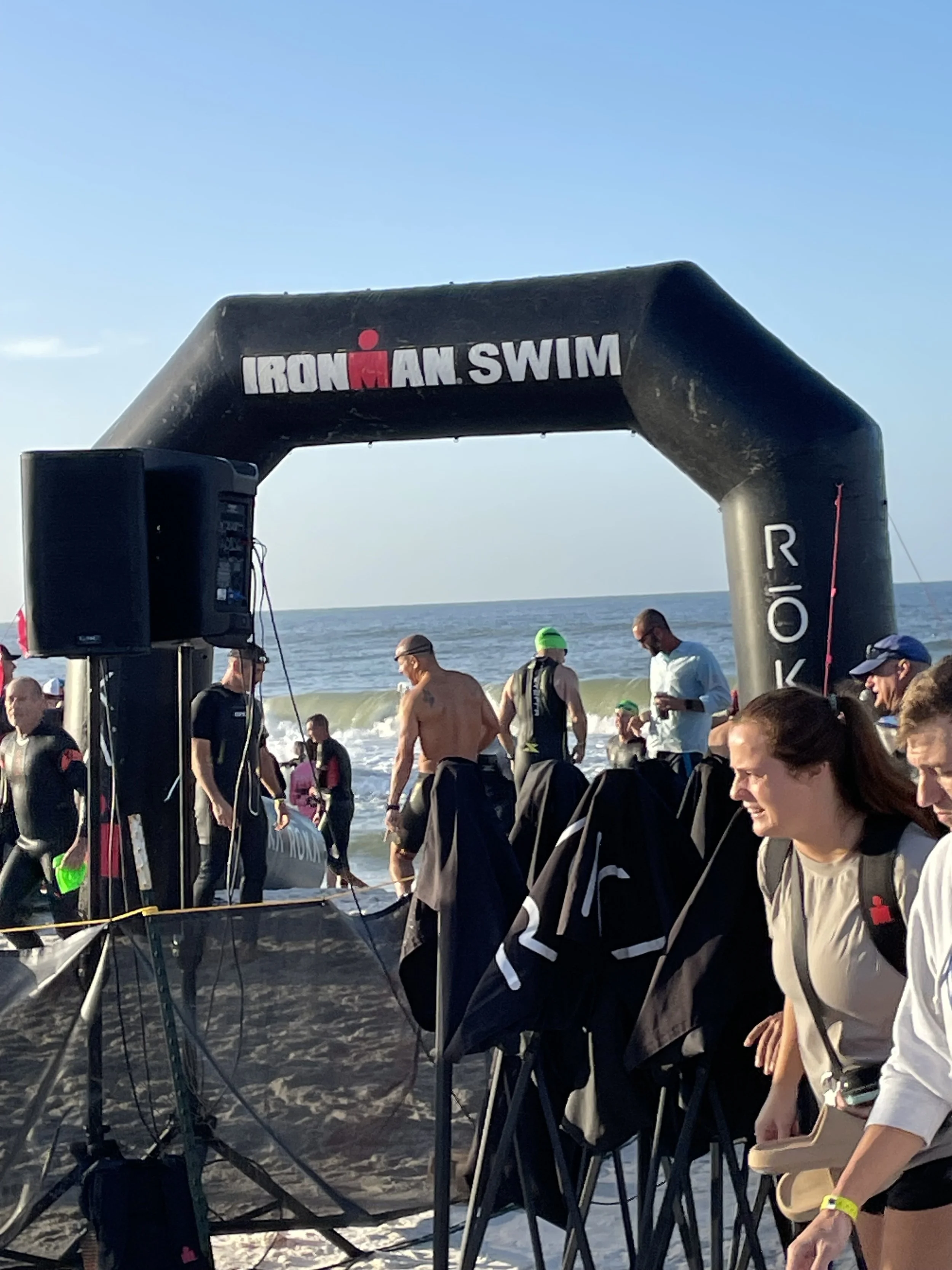 People gathered on the beach for the start of an Ironman swimming race, with an inflatable archway overhead and the ocean in the background.