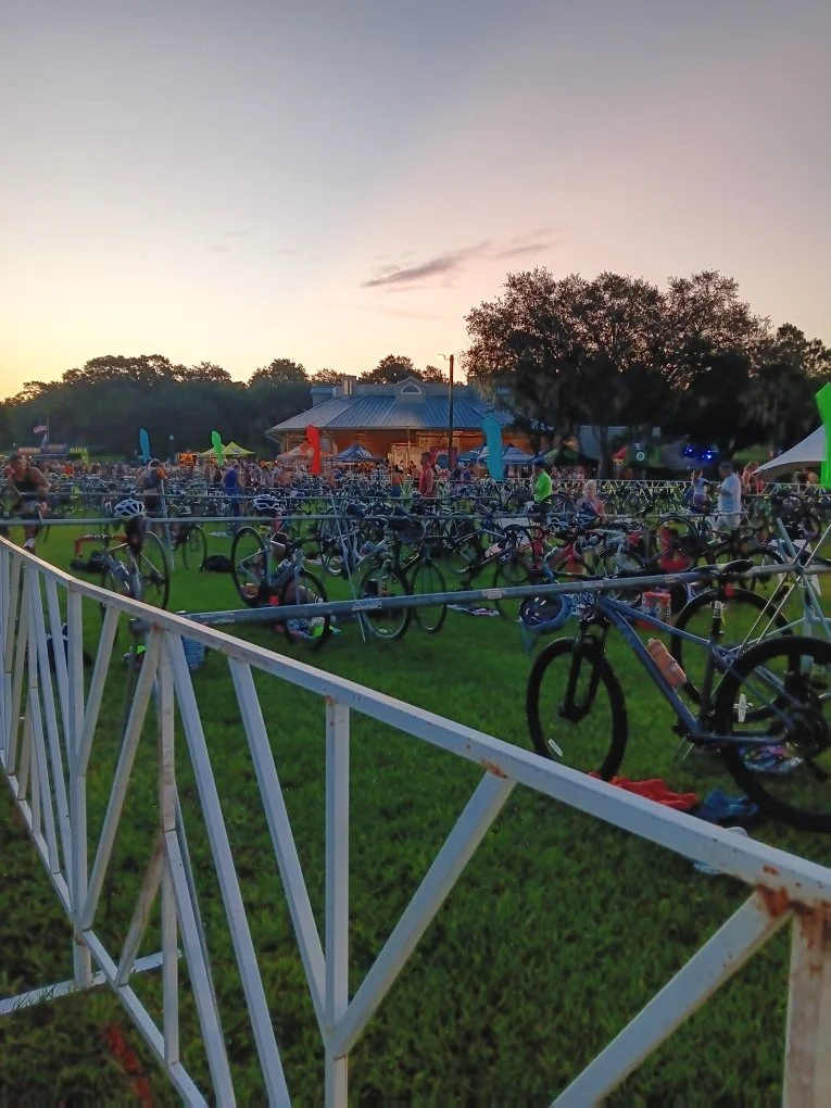 Outdoor event at dusk with numerous parked bicycles, a building with a tin roof, trees, and people gathered on a grassy area with colorful flags.