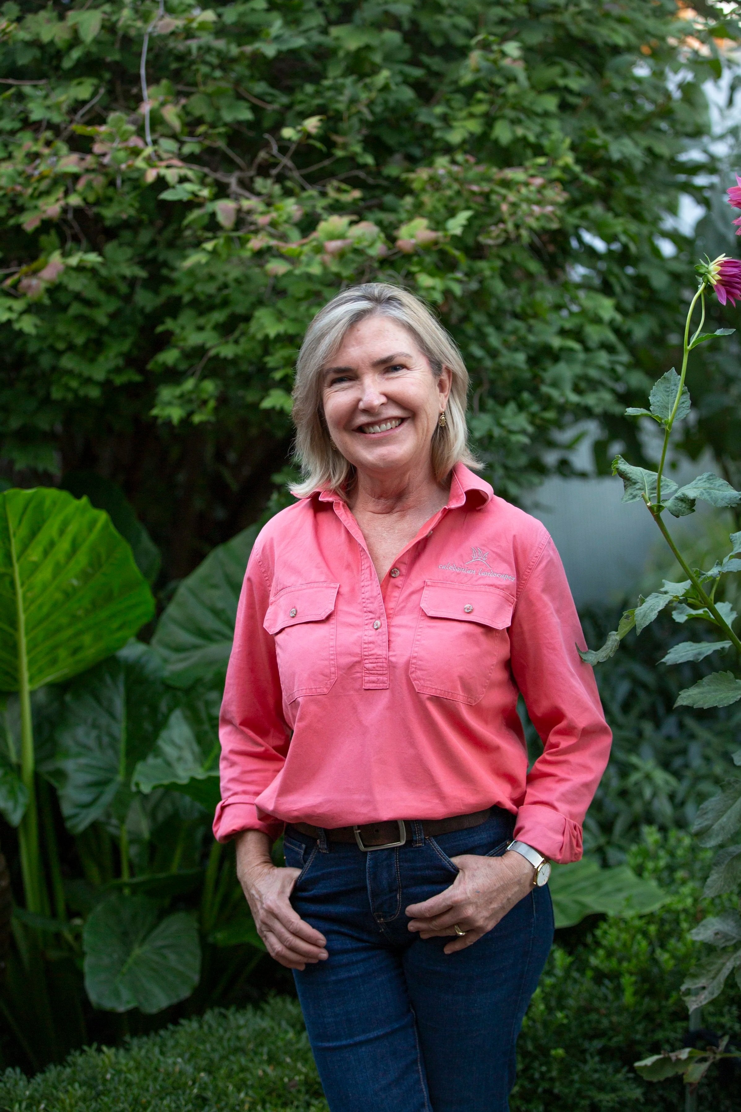 A smiling woman with gray hair wearing a pink shirt and blue jeans, standing outdoors in front of green foliage.