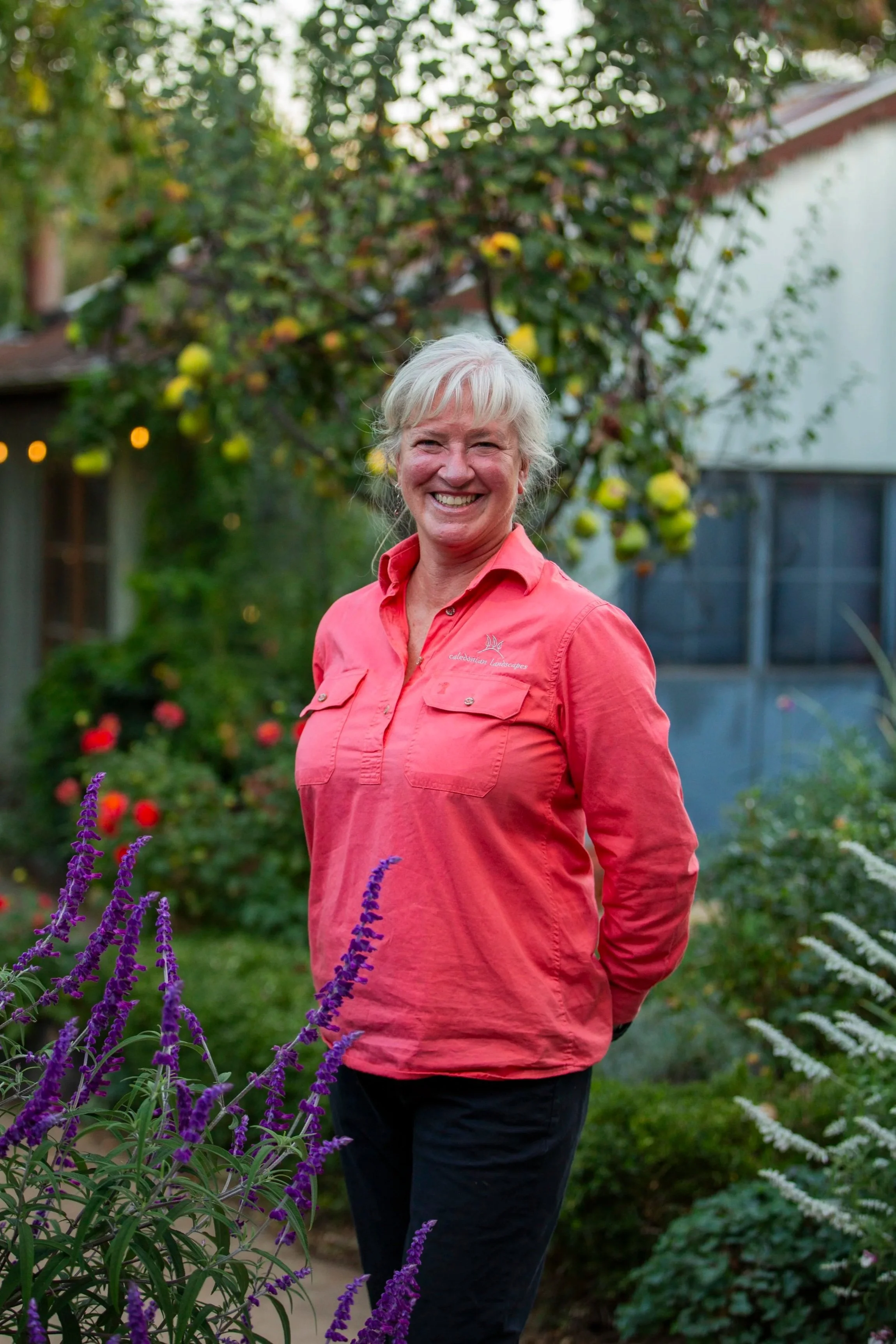 Smiling woman with short gray hair wearing a coral pink shirt standing in a garden with colorful flowers and green trees in the background.
